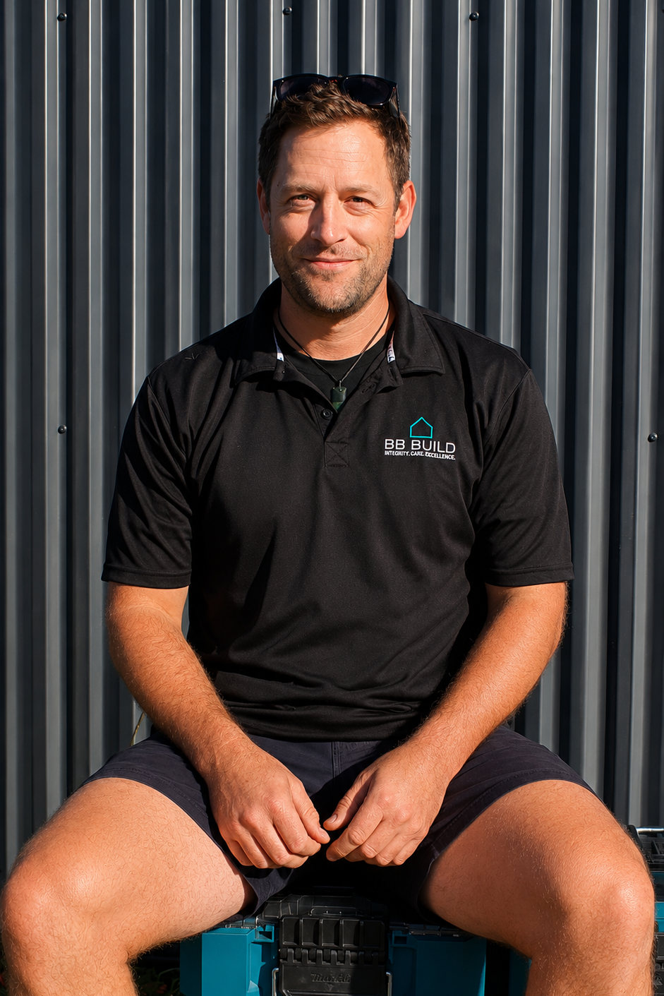 A man sitting outdoors in front of a metal wall, wearing a black polo shirt with a logo, sunglasses on his head, and shorts, looking at the camera.