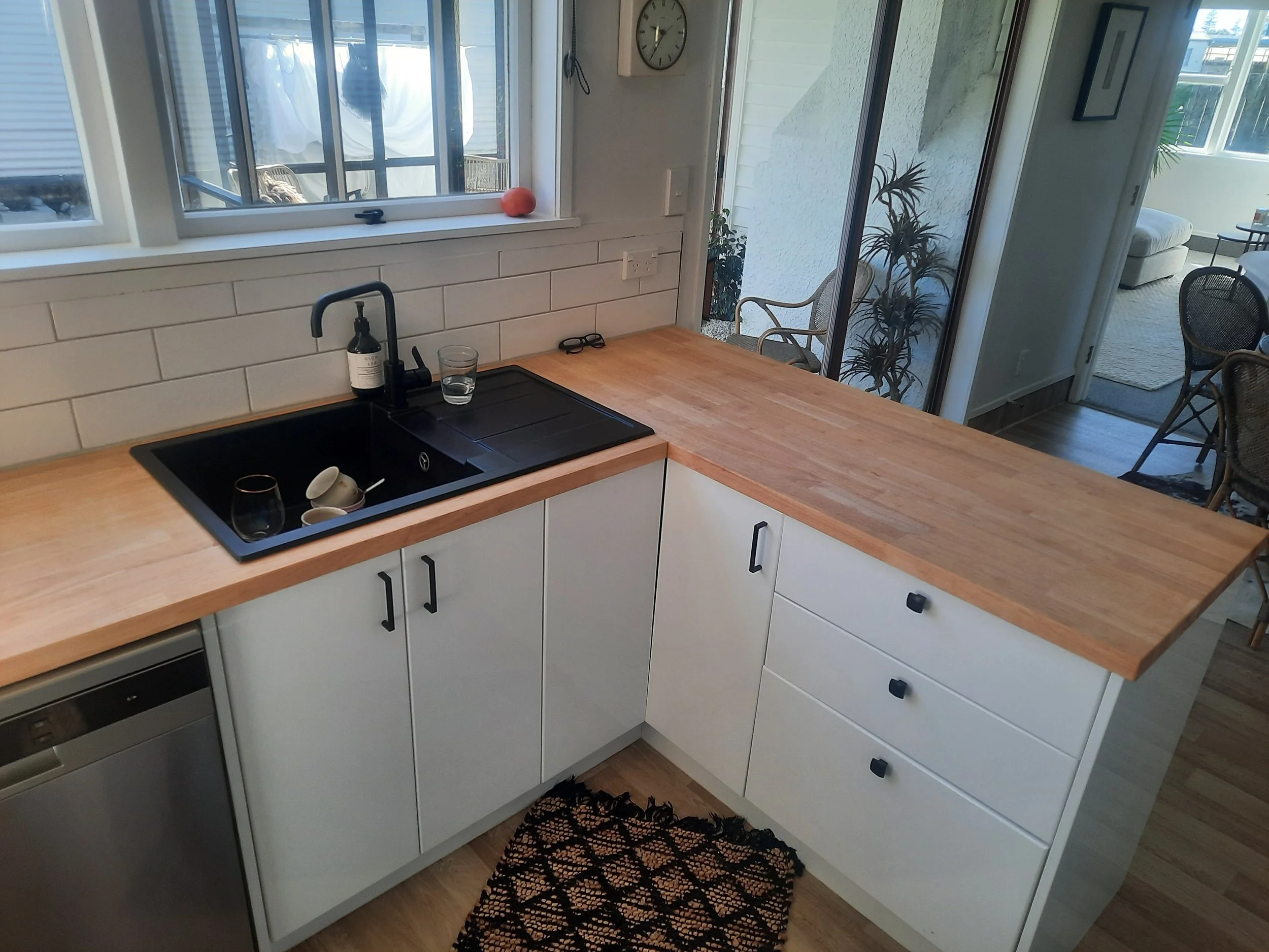 A kitchen with a wooden countertop, white cabinets, a black sink with dishes, a glass of water and soap dispenser, behind a window with a drying rack and a cat outside, a sliding door to a patio with potted plants and outdoor chairs, and a black fiber rug on a wooden floor.