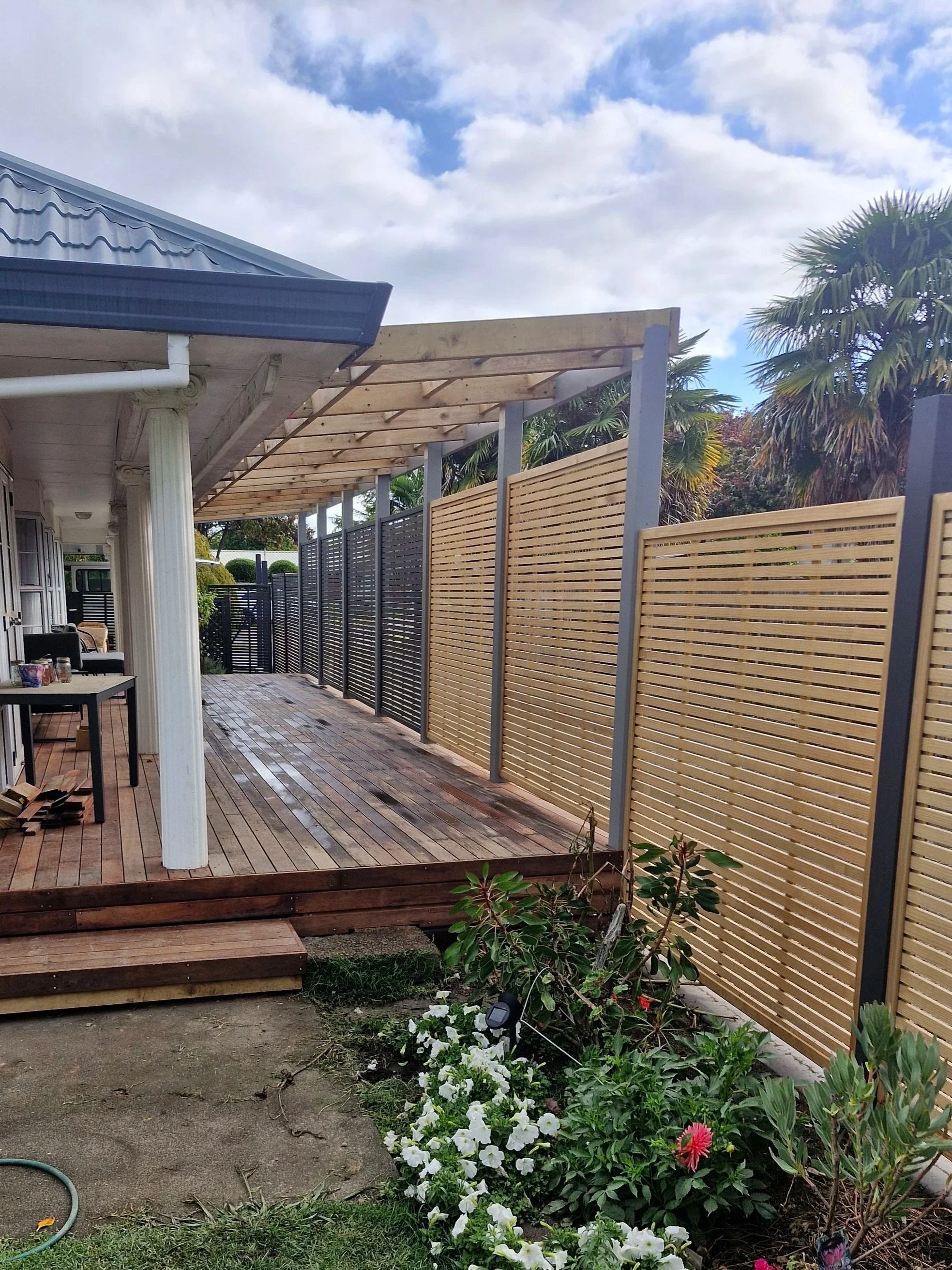 A backyard patio area with wooden decking, a newly installed wooden pergola frame, a privacy fence with horizontal slats, lush green plants and flowers, a cloudy sky with patches of blue, and some construction materials and a garden hose on the ground.