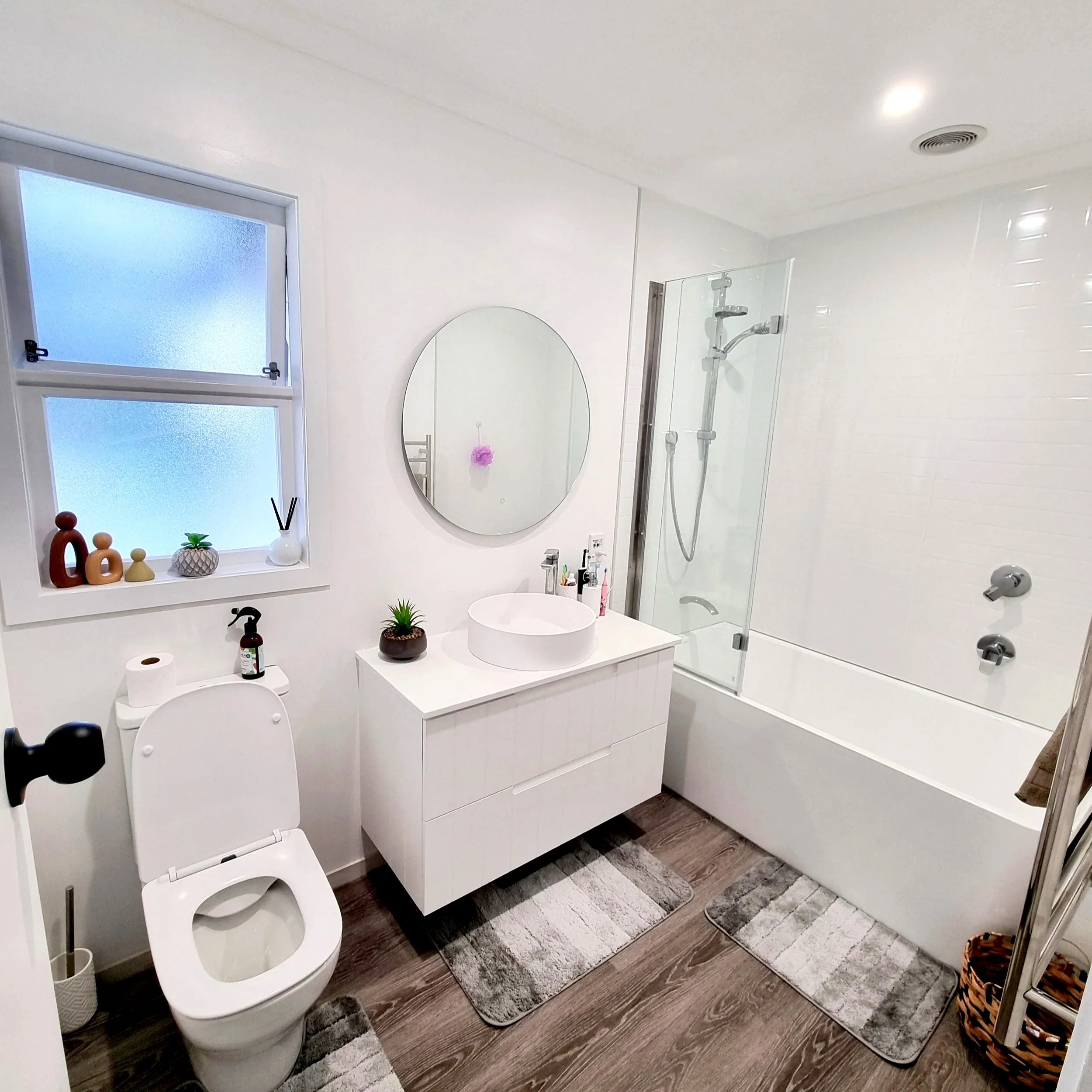 A modern bathroom with white walls and wooden flooring. Features a toilet, a white cabinet with a round vessel sink, a circular mirror, and a glass-enclosed shower with a showerhead. Decor includes wood-tone baskets, small potted plant, and decorative vases on the window sill.