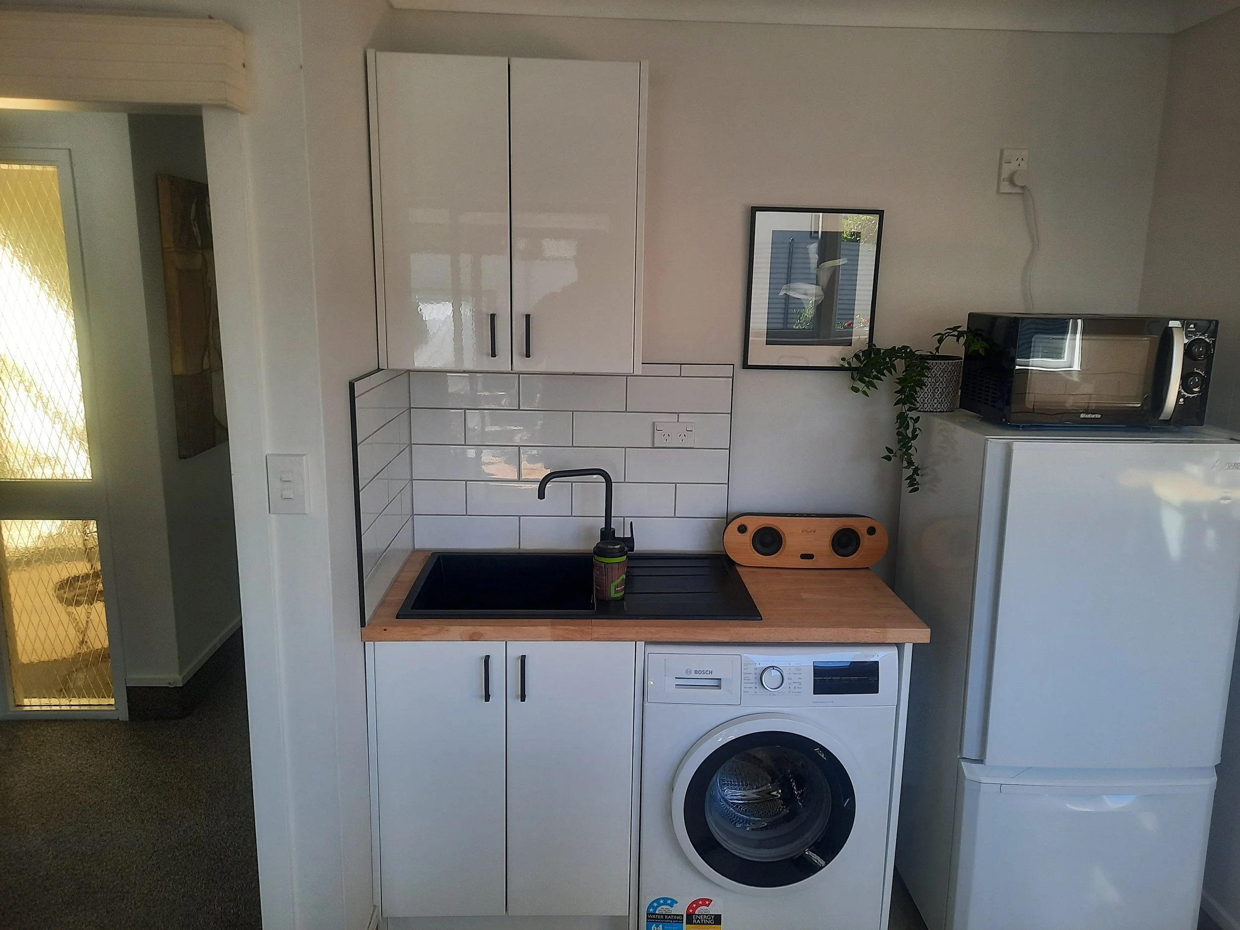 A small kitchen area with a white cabinet, a mirror, a microwave oven, a black sink with a faucet, a small wooden speaker, and a washing machine.