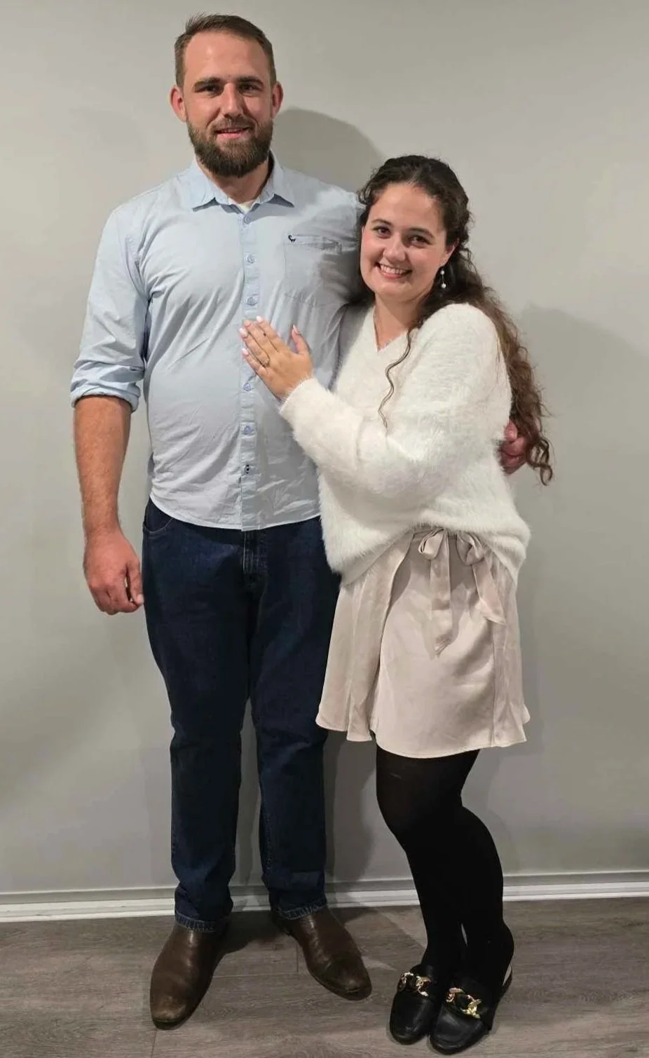 A man and woman standing together indoors, smiling at the camera. The woman has her hand on the man's chest displaying an engagement ring.