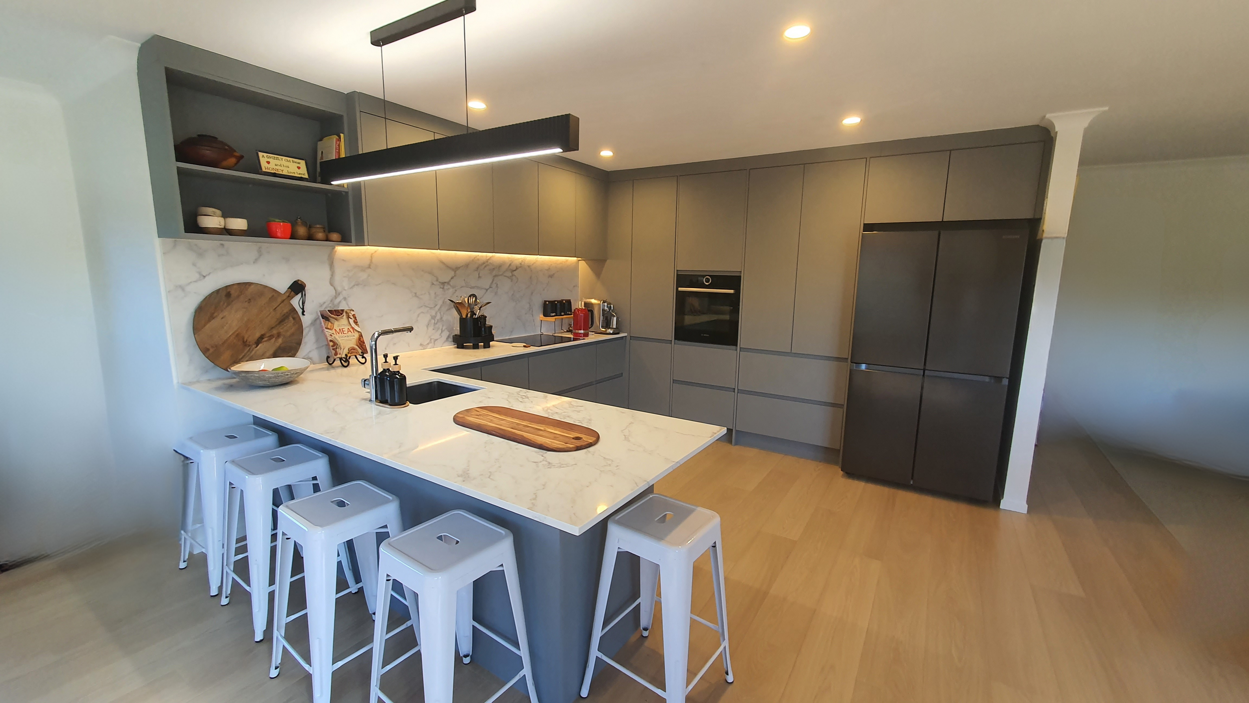 Modern kitchen with marble island, gray cabinets, open shelving with dishes and decor, black light fixture, and built-in appliances.