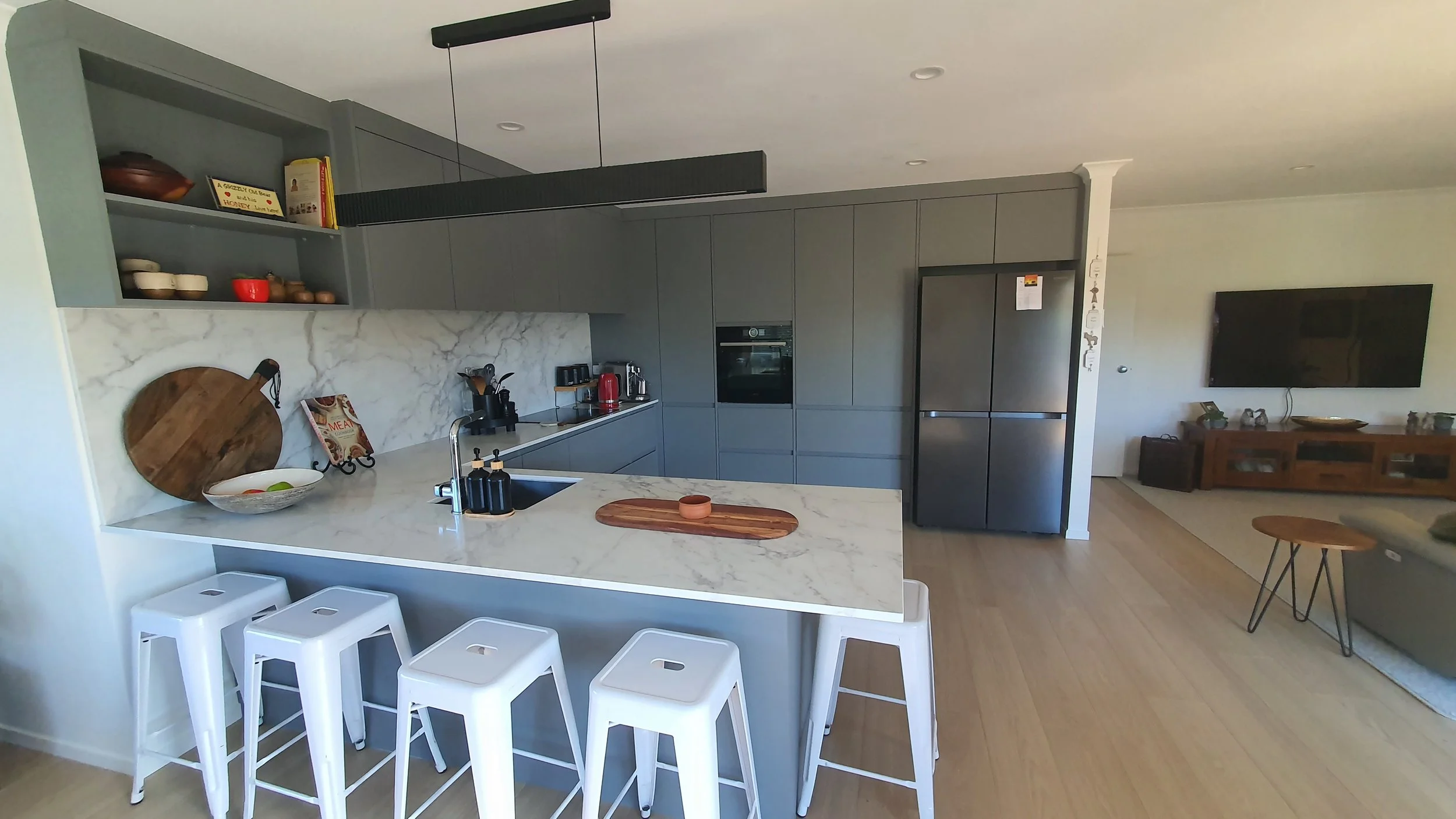 Modern kitchen with gray cabinetry, white marble island with a wooden cutting board, open shelves with bowls, a bowl of fruit, and a sink, adjacent to a living area with a TV and wooden furniture.