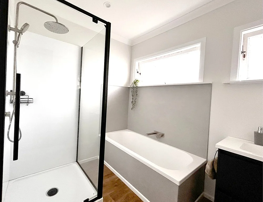 Modern bathroom with a glass shower enclosure, a white bathtub, a small black vanity, and two windows letting in natural light.