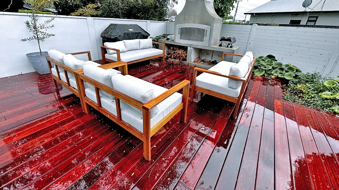 Rain-soaked red wooden deck with outdoor furniture, a white cushioned sofa set, plants in pots, and a built-in outdoor fireplace.