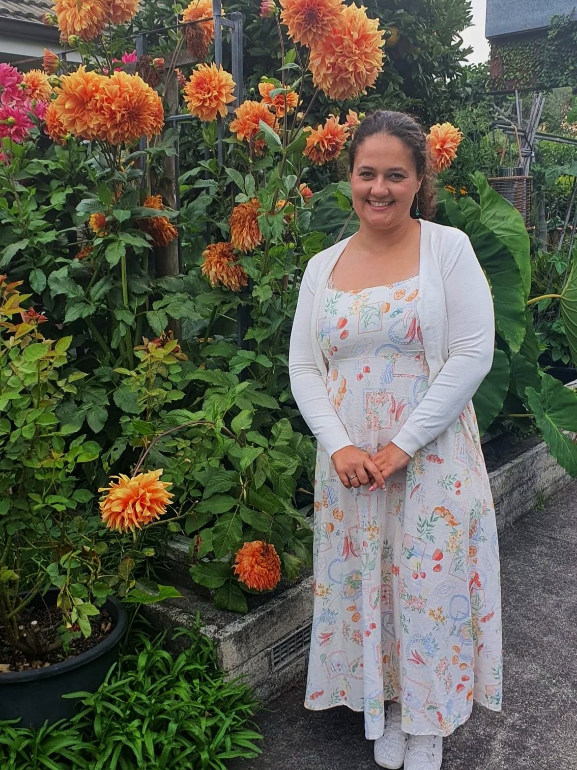 A woman smiling and standing in front of a lush garden filled with orange, pink, and red flowers, wearing a white cardigan over a colorful dress with fruit and floral patterns, on a sidewalk.
