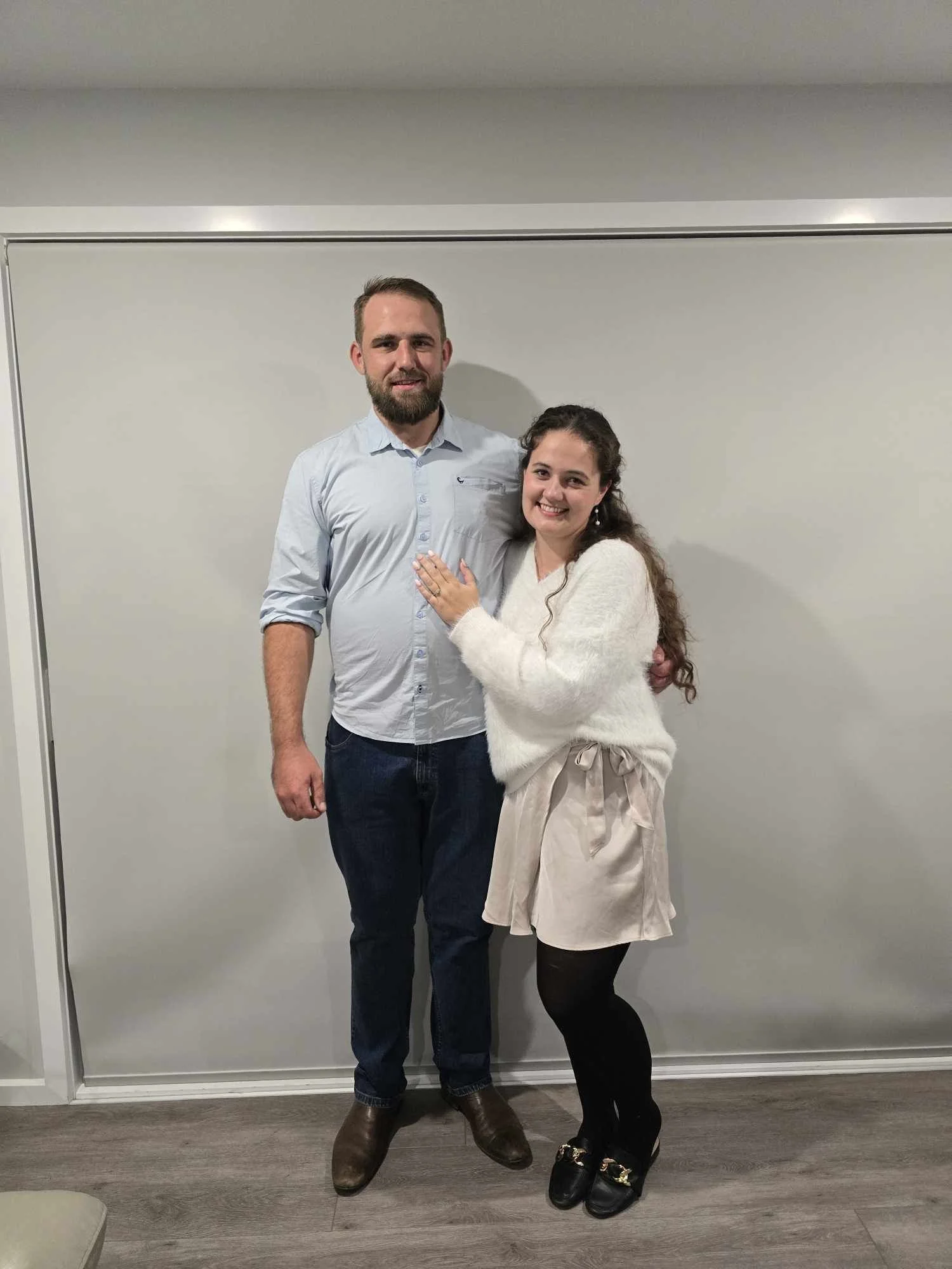 A man and woman standing together indoors in front of a plain white blank wall, smiling at the camera. The man is wearing a light blue button-up shirt, dark jeans, and brown boots. The woman is wearing a white fuzzy sweater, a beige skirt with a bow, black tights, and black shoes with gold accents. They are standing close, with their bodies slightly angled towards each other.
