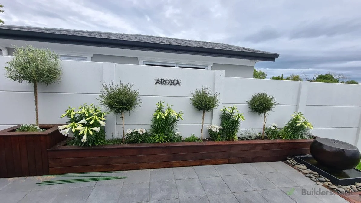 A modern backyard garden with a white wall, a long wooden planter box filled with plants including white lilies, three small trees, and a black water fountain with smooth stones surrounding it, under a cloudy sky.