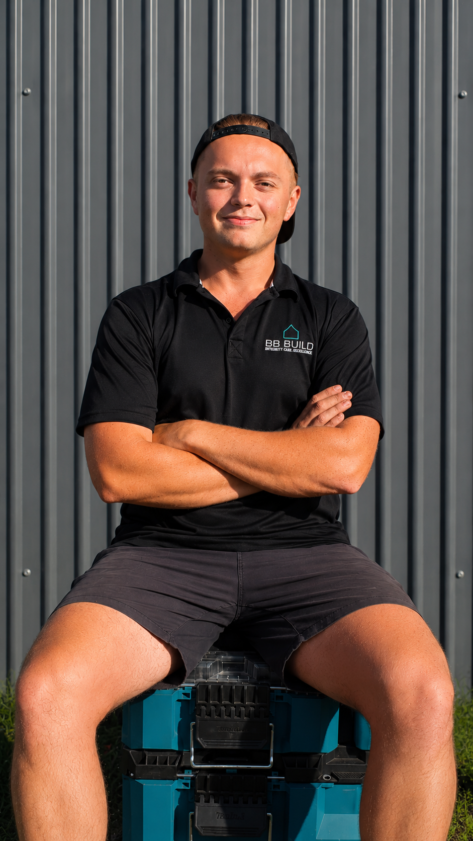 Young man sitting on a toolbox in front of a metal wall, wearing a black polo shirt with 'BB Build' logo, black shorts, and a backward cap.