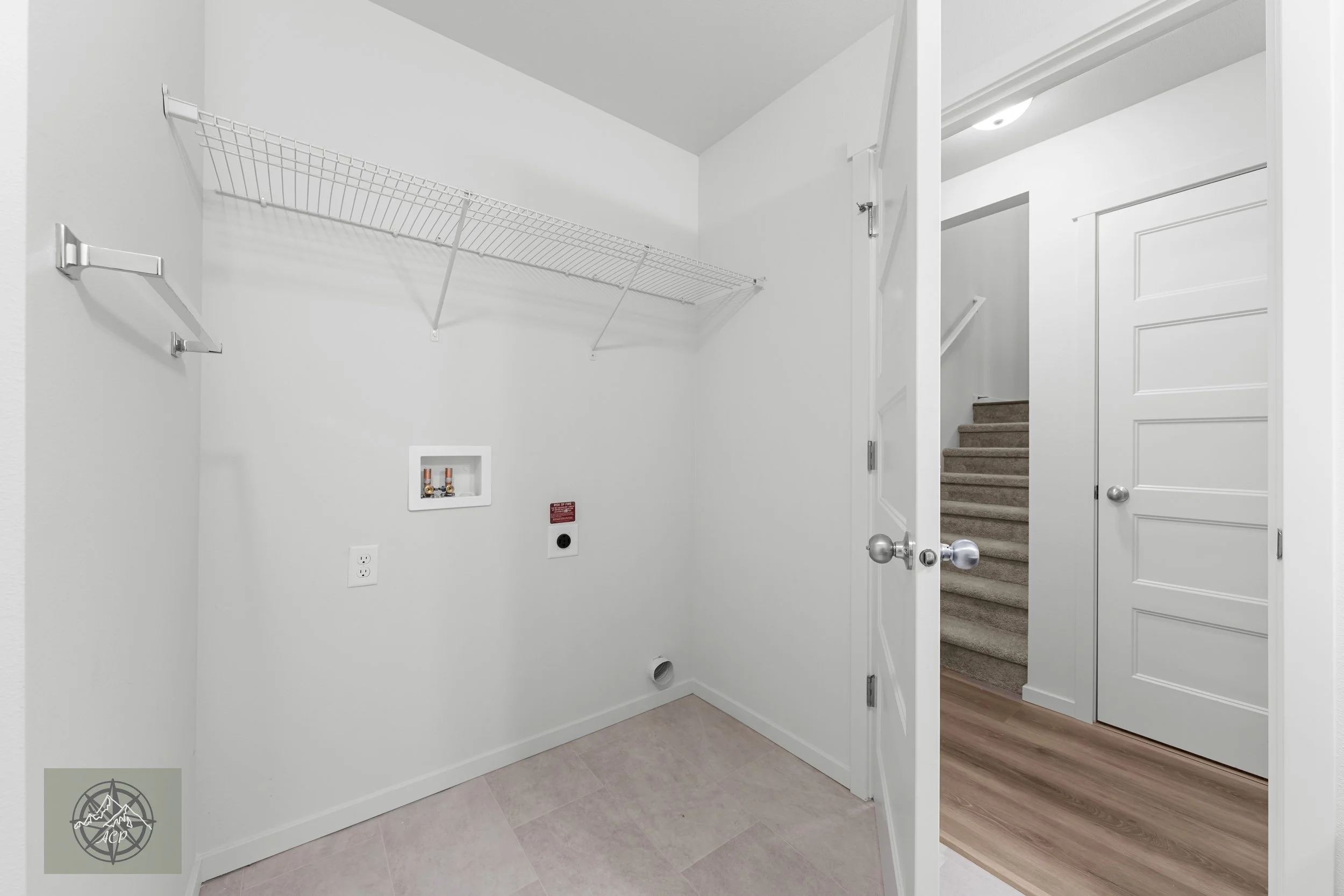 Empty laundry room with white walls, wire shelf, and hookups for washer and dryer, viewed through an open door with stairs outside.