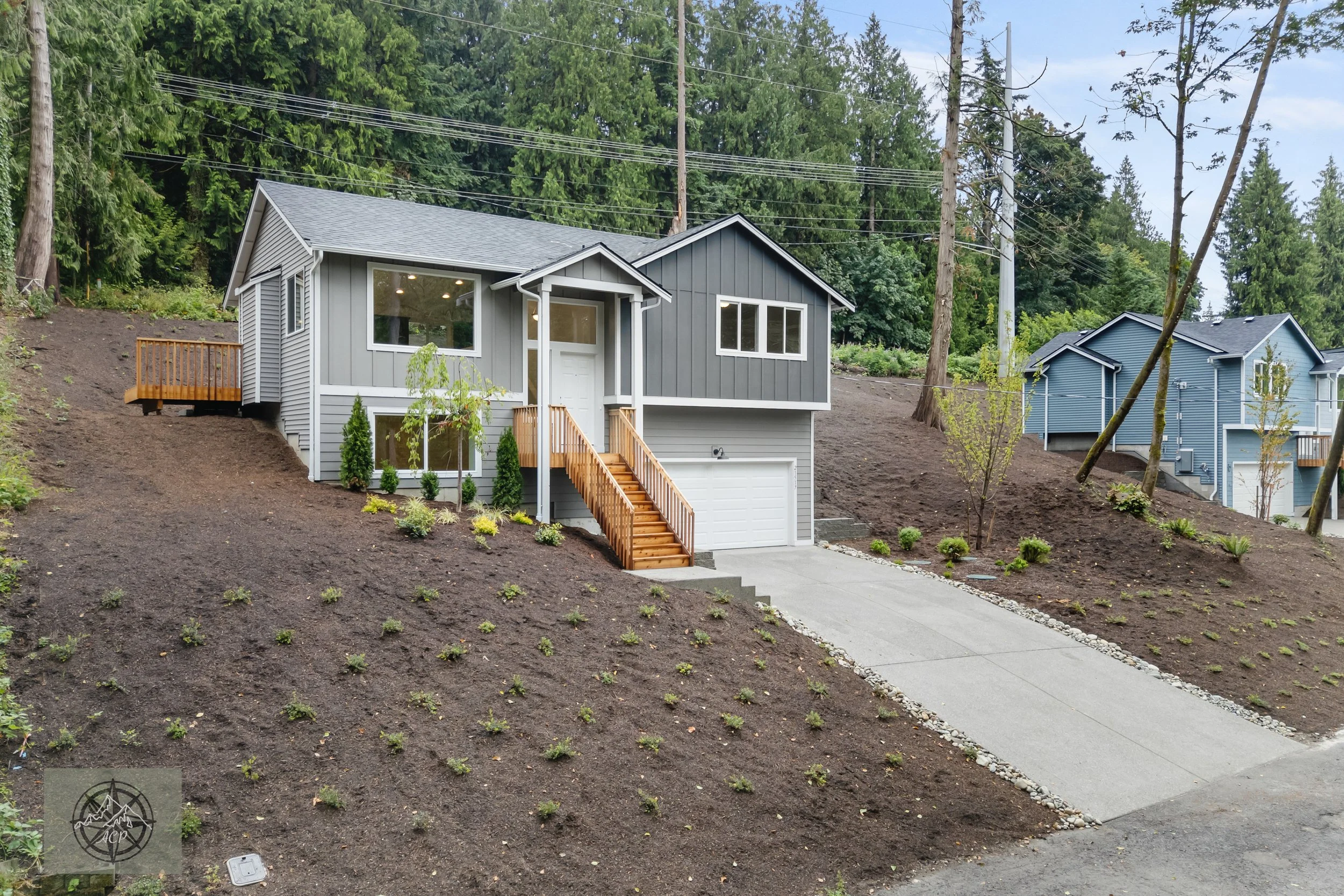 Newly built gray and dark gray house on a sloped lot with a driveway and landscaped front yard, surrounded by tall trees.
