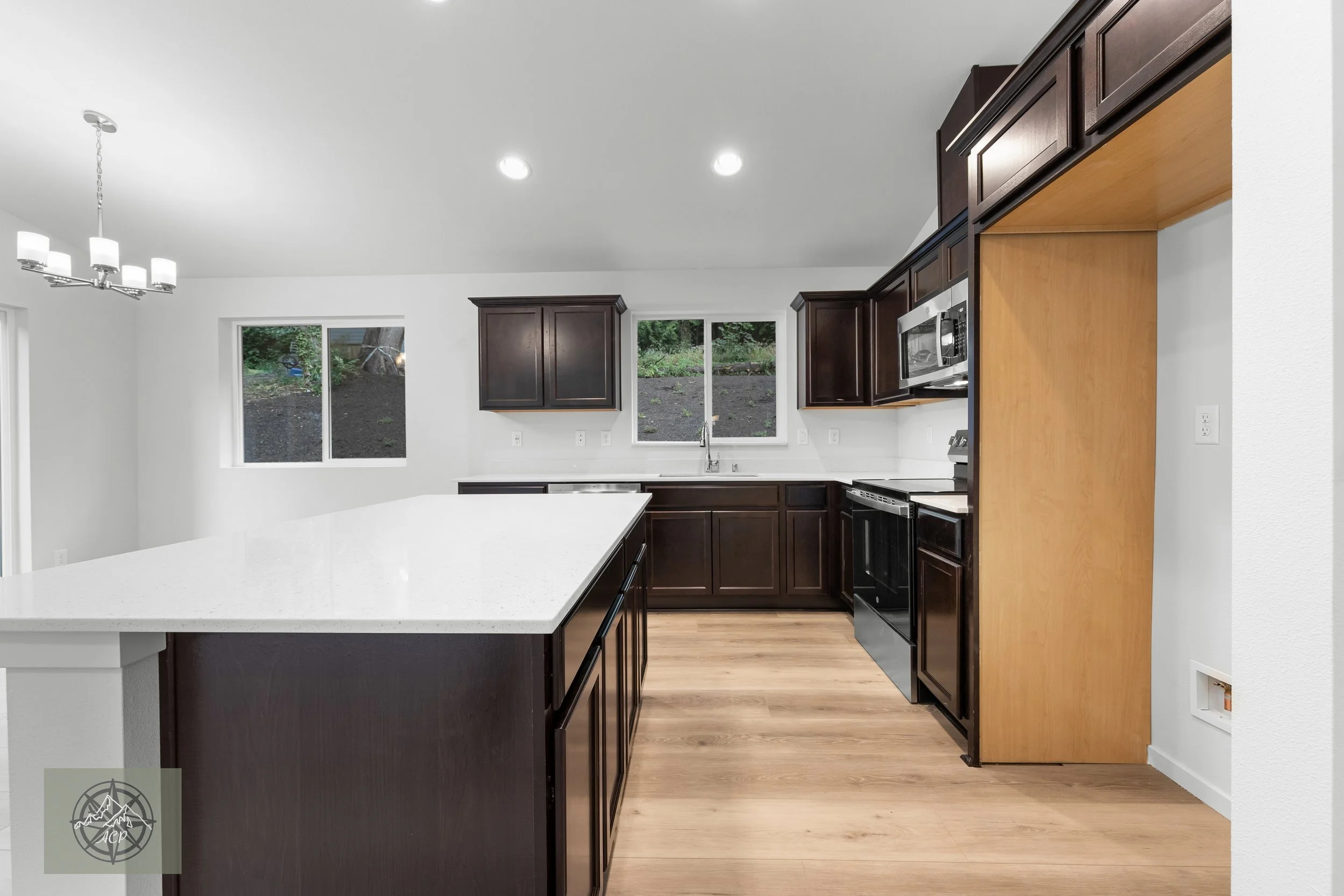 Modern kitchen with white countertops, dark brown cabinets, and stainless steel appliances. Hardwood flooring and two windows overlooking greenery.