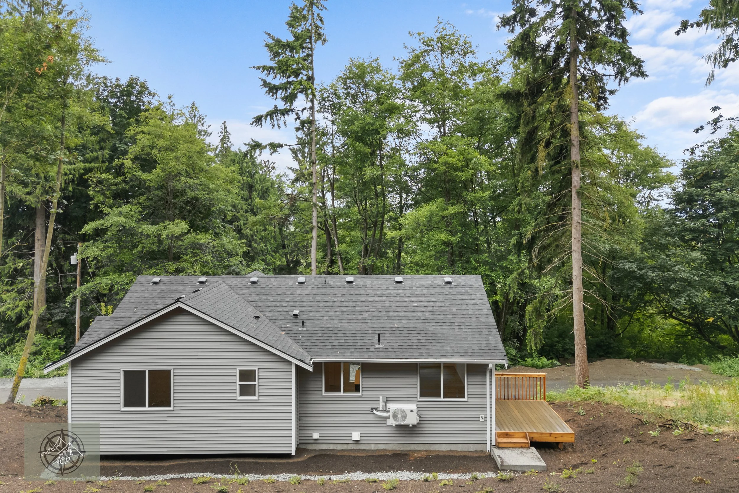 New gray house with a gable roof and a small wooden deck, surrounded by green trees and a dirt yard.