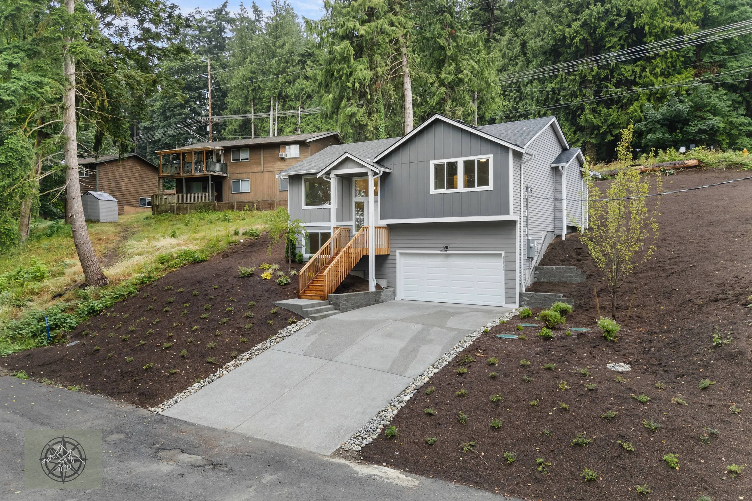New gray two-story house with a white garage door and wooden stairs, situated on a sloped lot with freshly landscaped yard and young plants, in a wooded area with tall trees and neighboring houses.