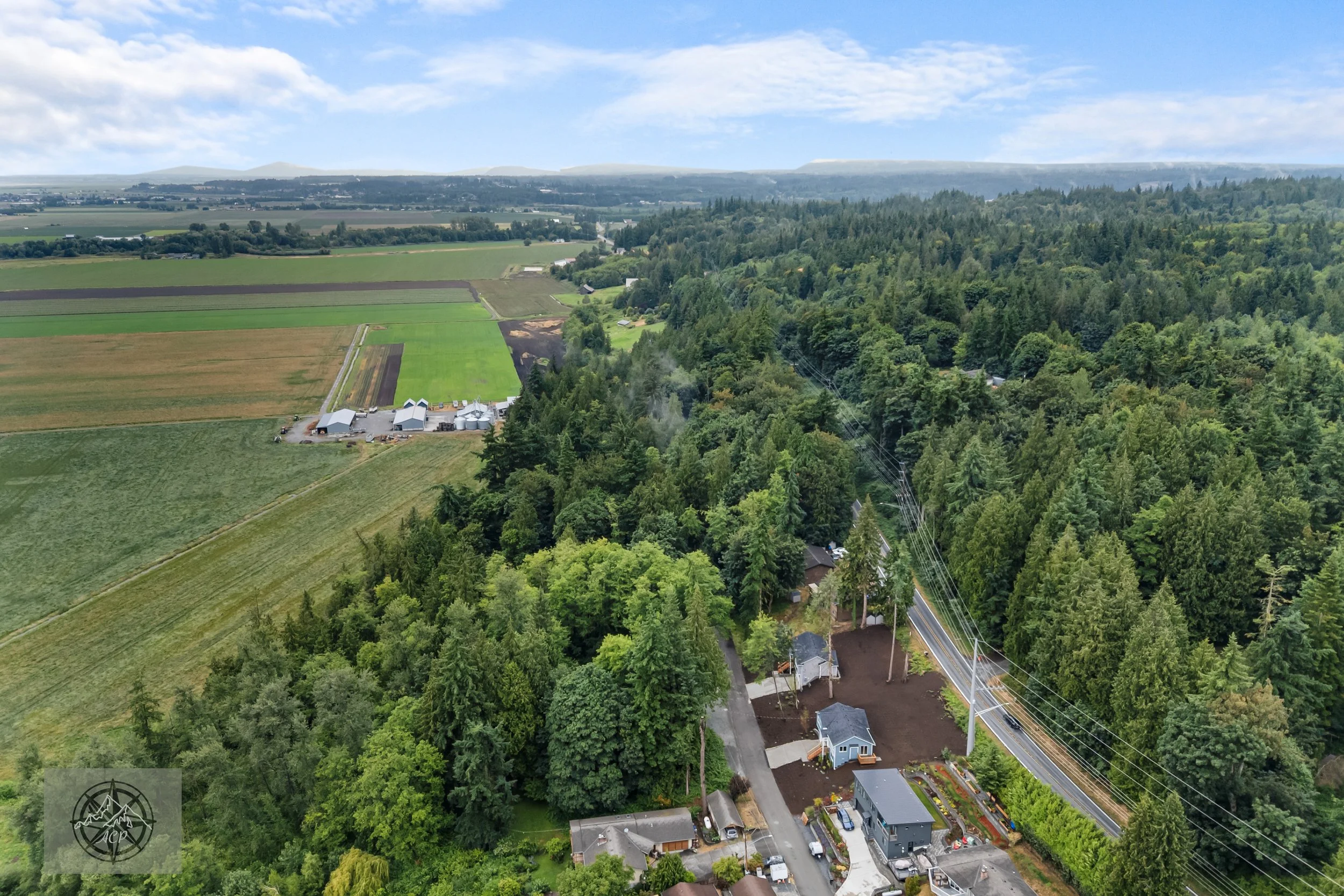 Aerial view of farmland, residential houses, and a forested area in the countryside with mountains in the background, under a partly cloudy sky.
