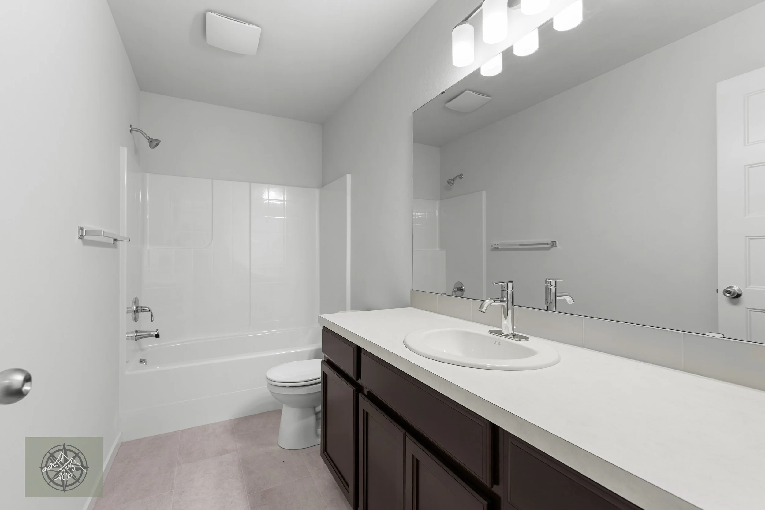 A bathroom with a white bathtub, toilet, dark wood vanity with a white countertop, and a large mirror. White walls and tile surround the bathtub, with ceiling-mounted lights and a vent.
