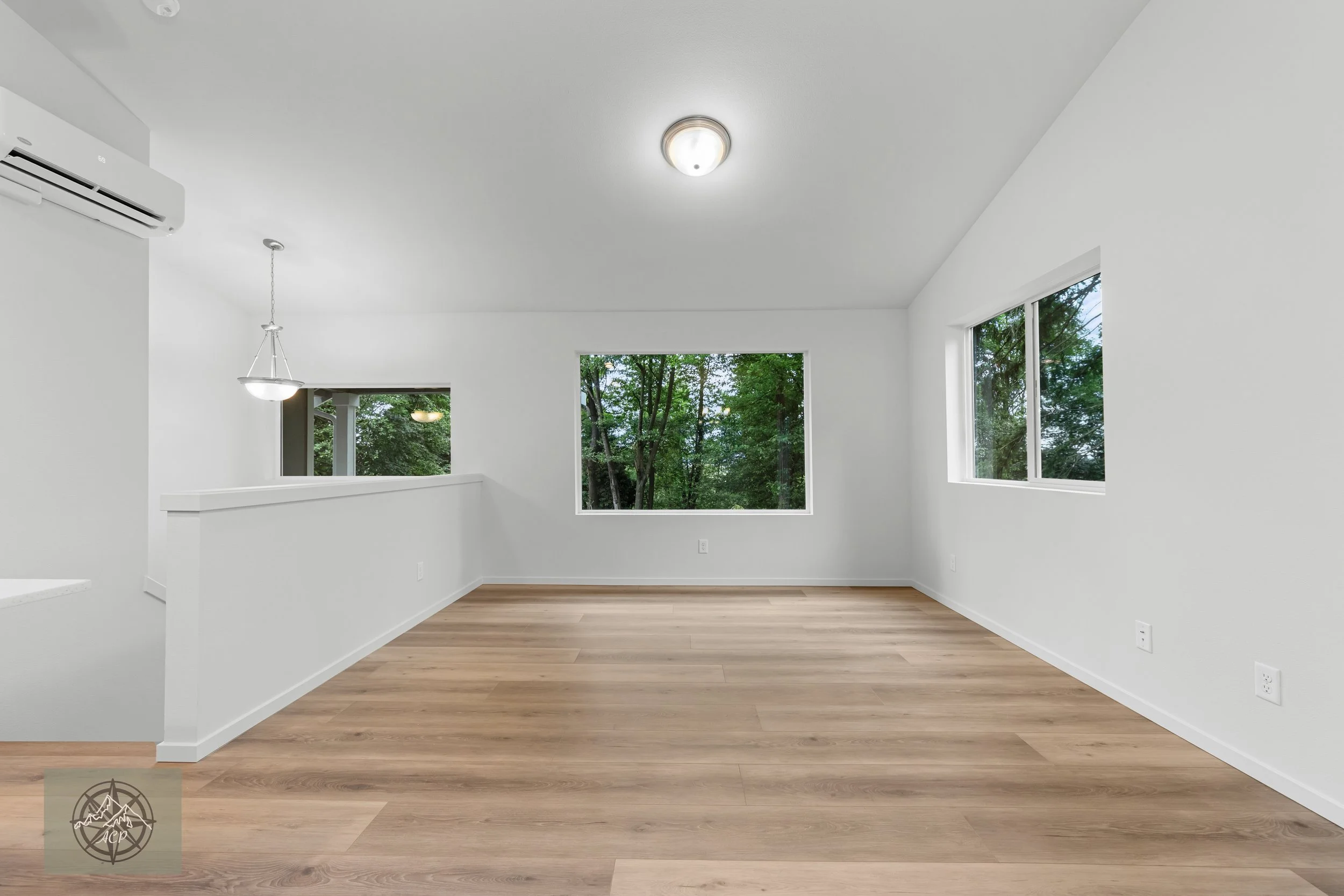 Empty living room with wood flooring, white walls, multiple windows showing outdoor trees, ceiling lights, and an air conditioner