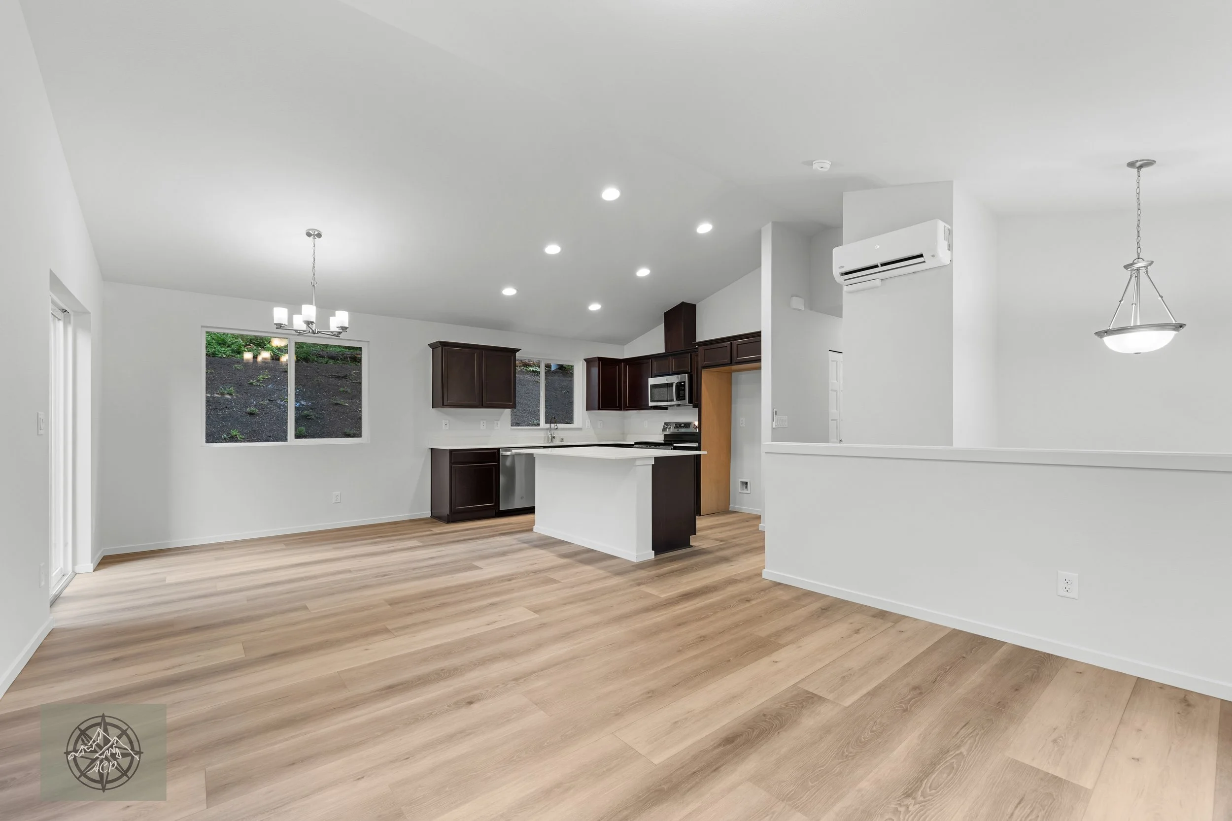Empty modern open-plan kitchen and dining area with white walls, wooden flooring, dark brown cabinets, stainless steel appliances, and two hanging light fixtures.