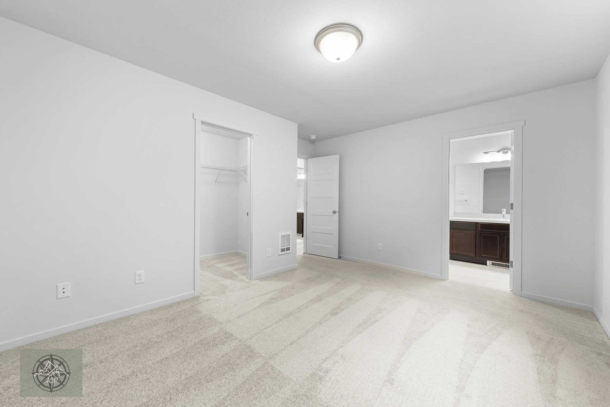 Empty bedroom with beige carpet, white walls, two doorways leading to a closet and a bathroom, ceiling light, and a vent on the wall.