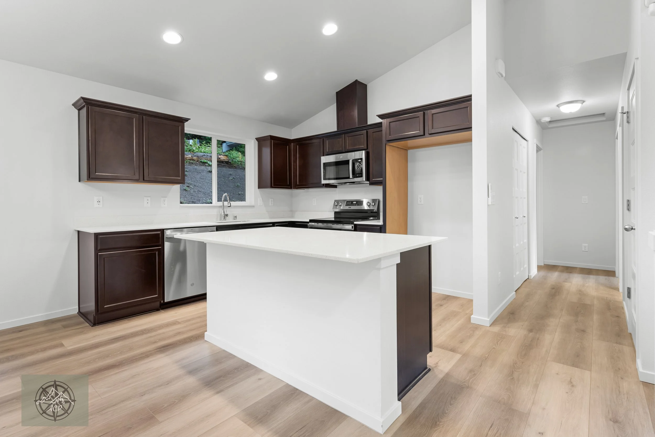 Modern kitchen with dark wood cabinets, white countertops, stainless steel appliances, and a large window over the sink.