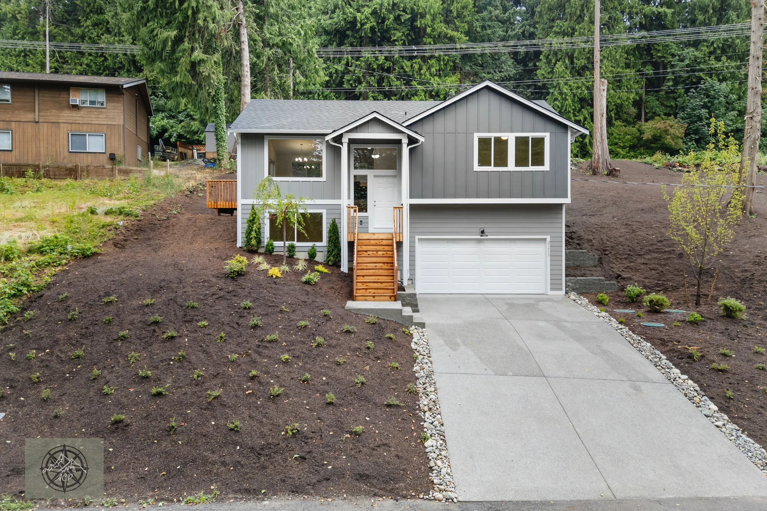 Front view of a newly built two-story house with gray siding, attached garage, concrete driveway, and small landscaped yard with newly planted shrubs and young trees.