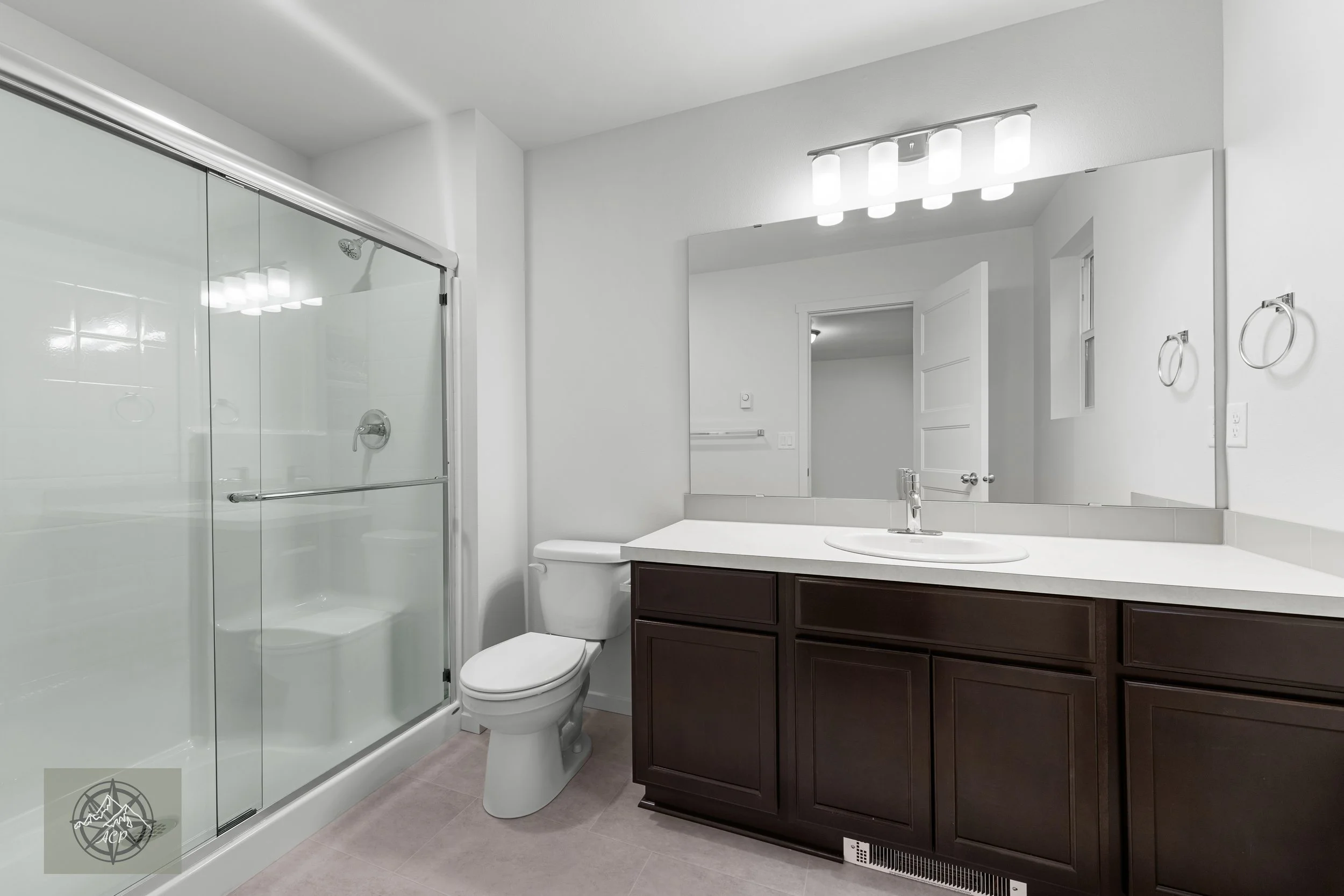 Modern bathroom with a glass-enclosed shower, white toilet, dark wood vanity with a white countertop, large mirror, and white walls.