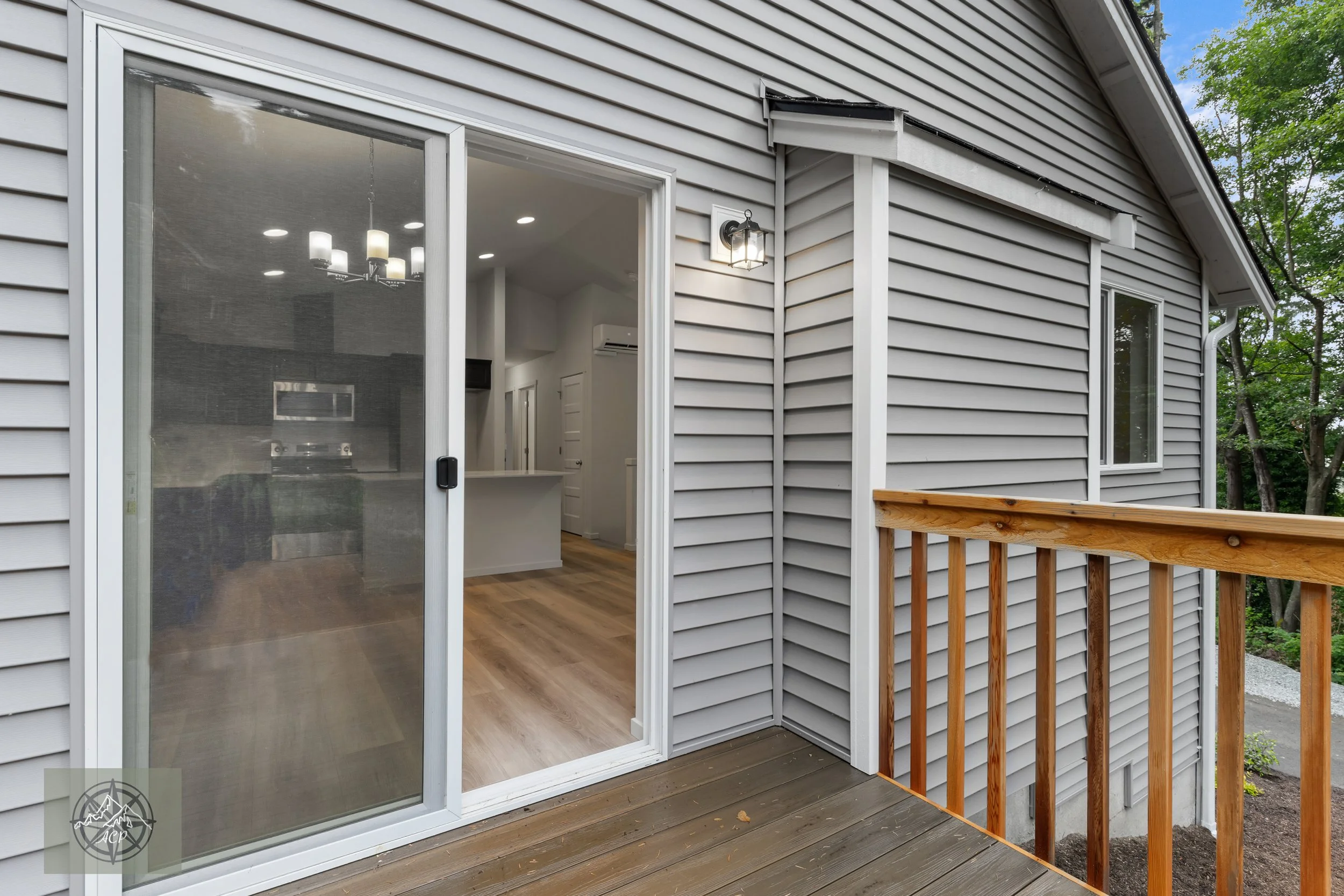 View of a house's backyard sliding glass door opening to a small wooden deck, with a view into the interior kitchen and living area. The house has grey vinyl siding, and a small outdoor wall-mounted light fixture is next to the door.