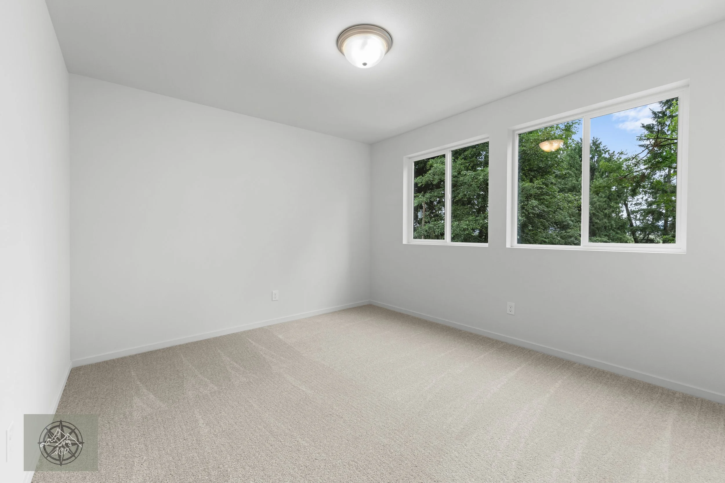 A clean, empty room with white walls, beige carpet, and three large windows with greenery outside, a ceiling light fixture, and white baseboards.