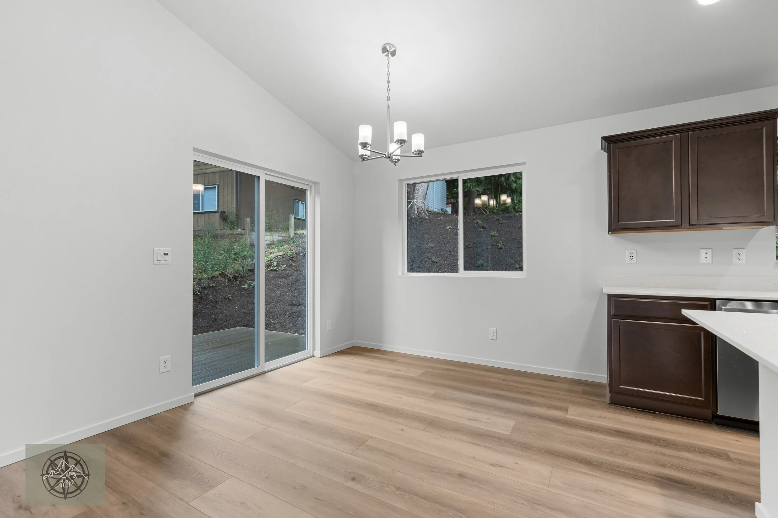Empty kitchen and dining area with wooden flooring, white walls, dark wood cabinets, a sliding glass door leading outside, a window overlooking a backyard hillside, and a modern chandelier.