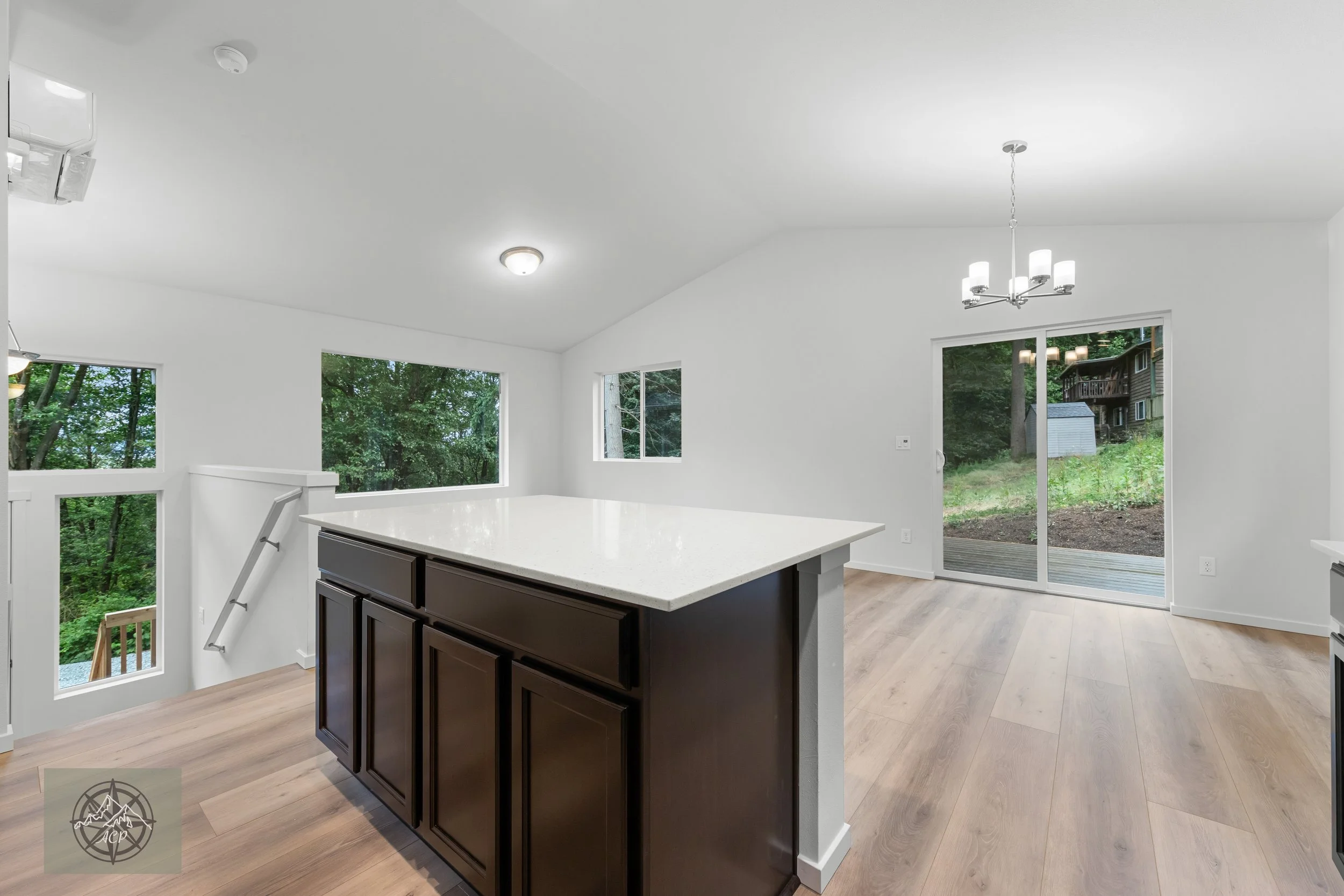 Empty modern kitchen with white walls, dark cabinets, light wood flooring, large windows, a sliding glass door, and a dining chandelier.