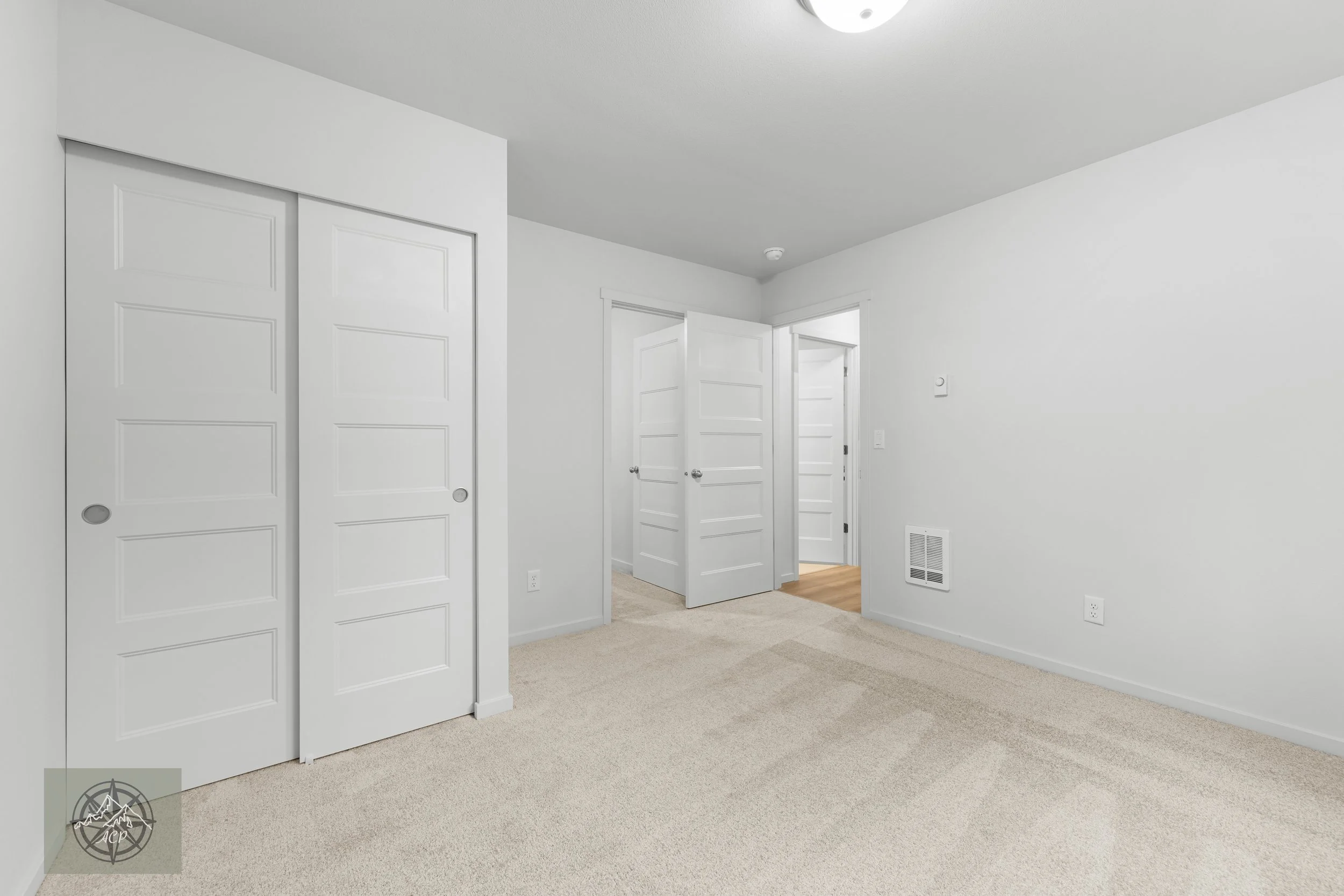 Empty bedroom with white walls, beige carpet, and multiple white doors, including closet doors and a doorway leading to another room.