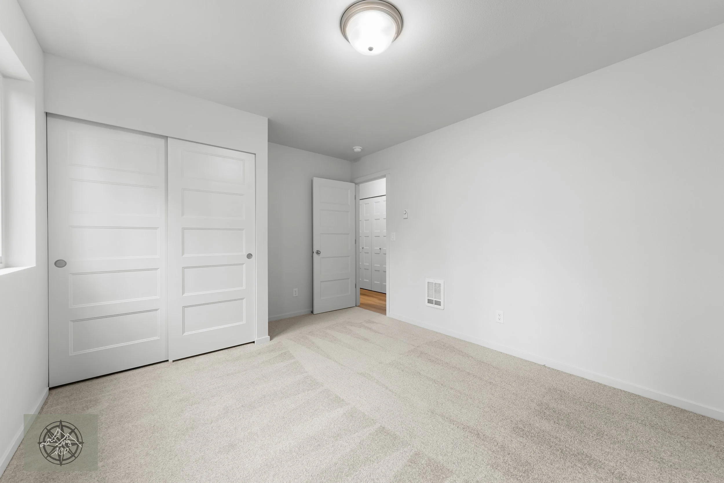 Empty bedroom with white walls, beige carpet, a closed closet with sliding doors, an open door revealing another room, and a ceiling light fixture.