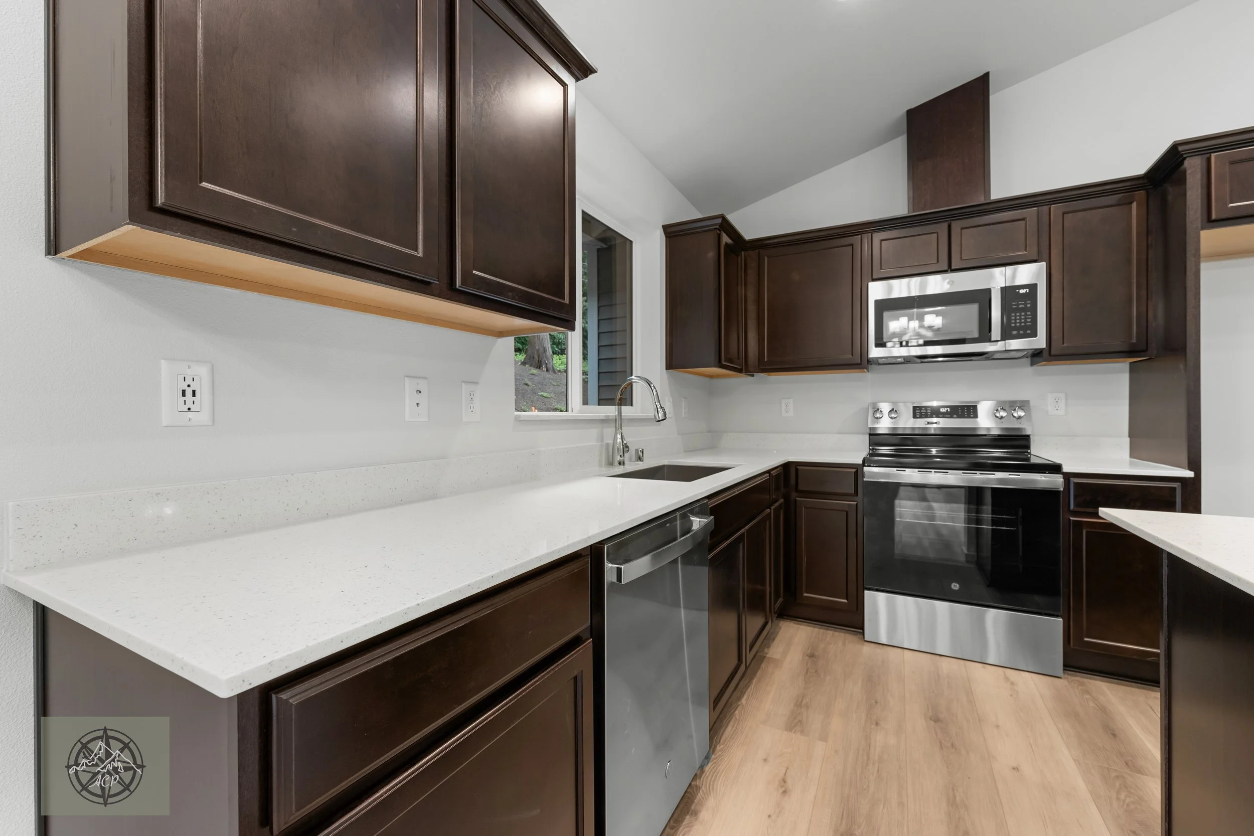 Modern kitchen with dark wood cabinets, white speckled countertops, stainless steel oven, microwave, and dishwasher, and a small window above the sink.