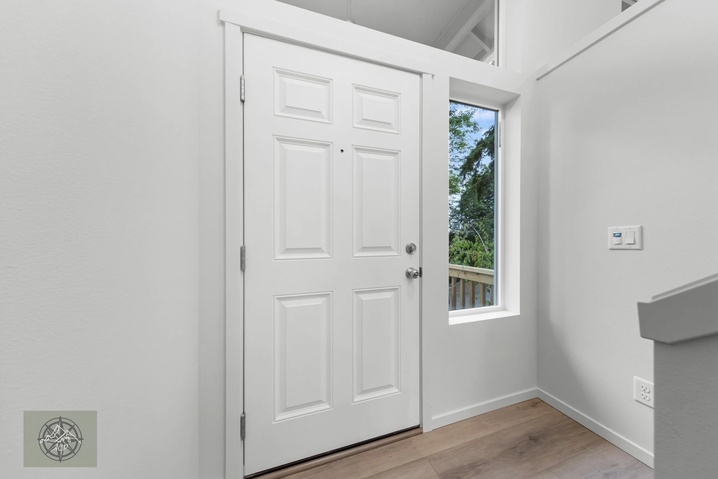 White front door with a half-window to the right, showing trees outside, inside a home with light-colored walls and wood flooring.