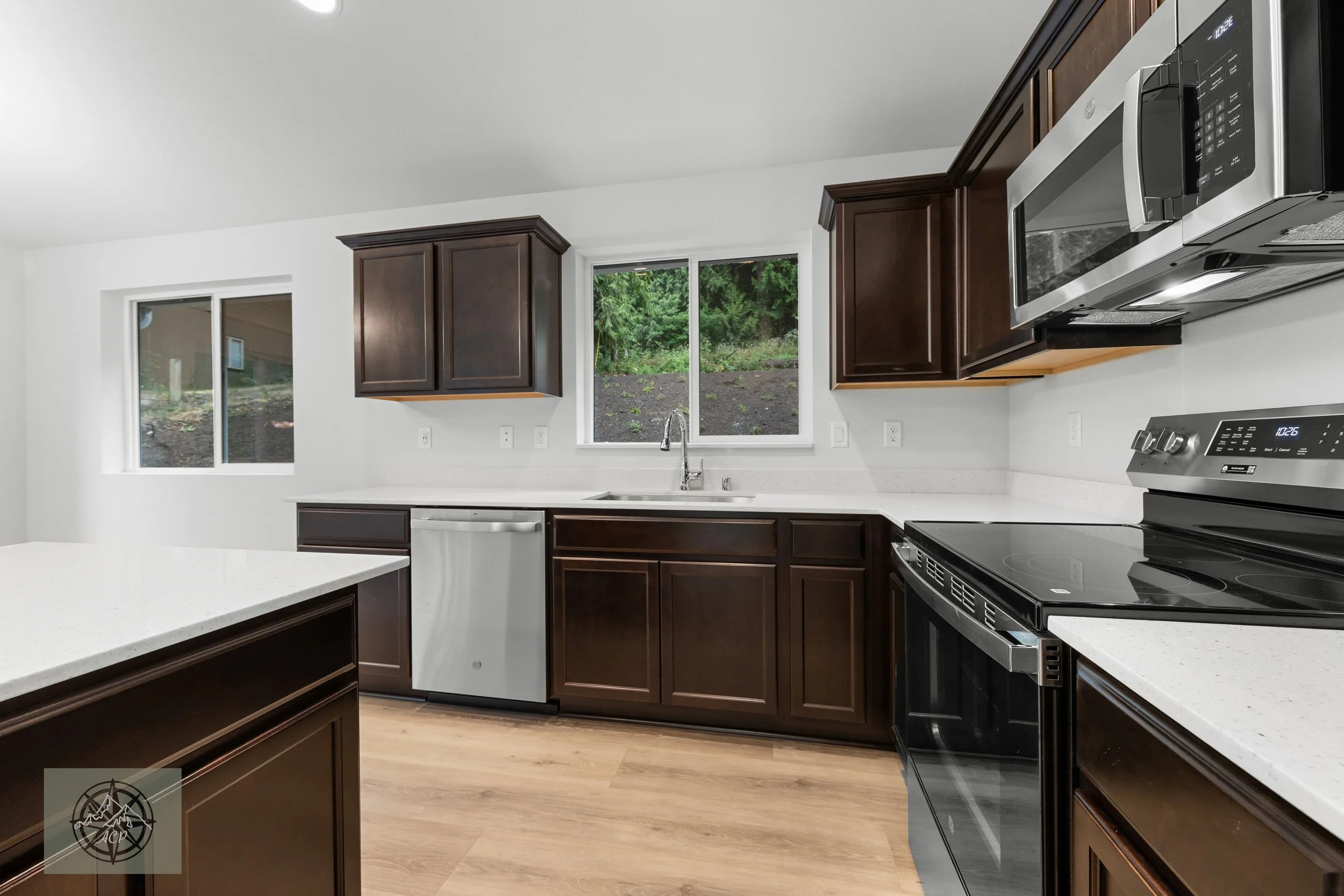 Clean modern kitchen with dark wooden cabinets, white countertops, stainless steel appliances, and two windows overlooking greenery.