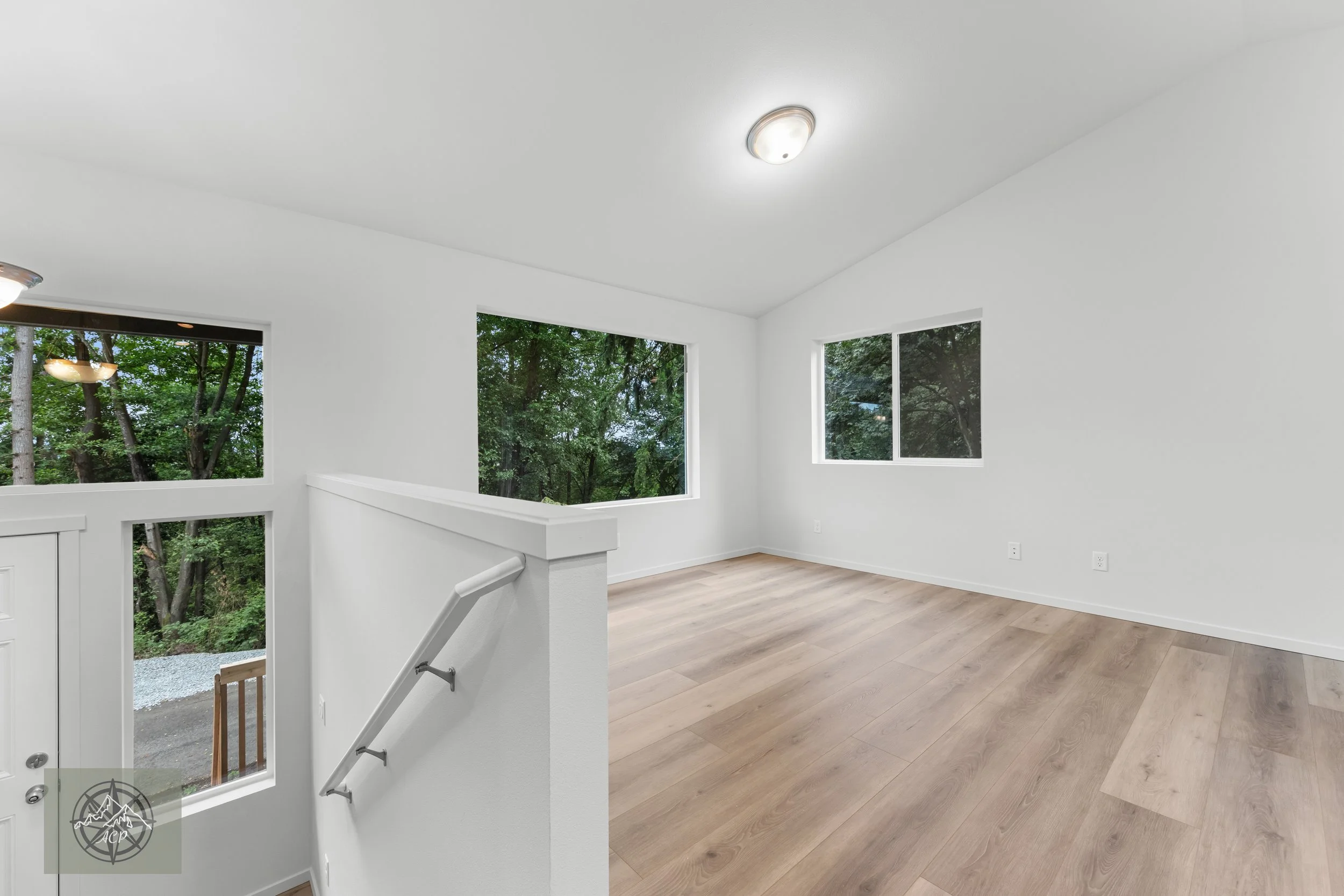 Empty, bright room with white walls, large windows showing outdoor trees, wood flooring, and ceiling lights.
