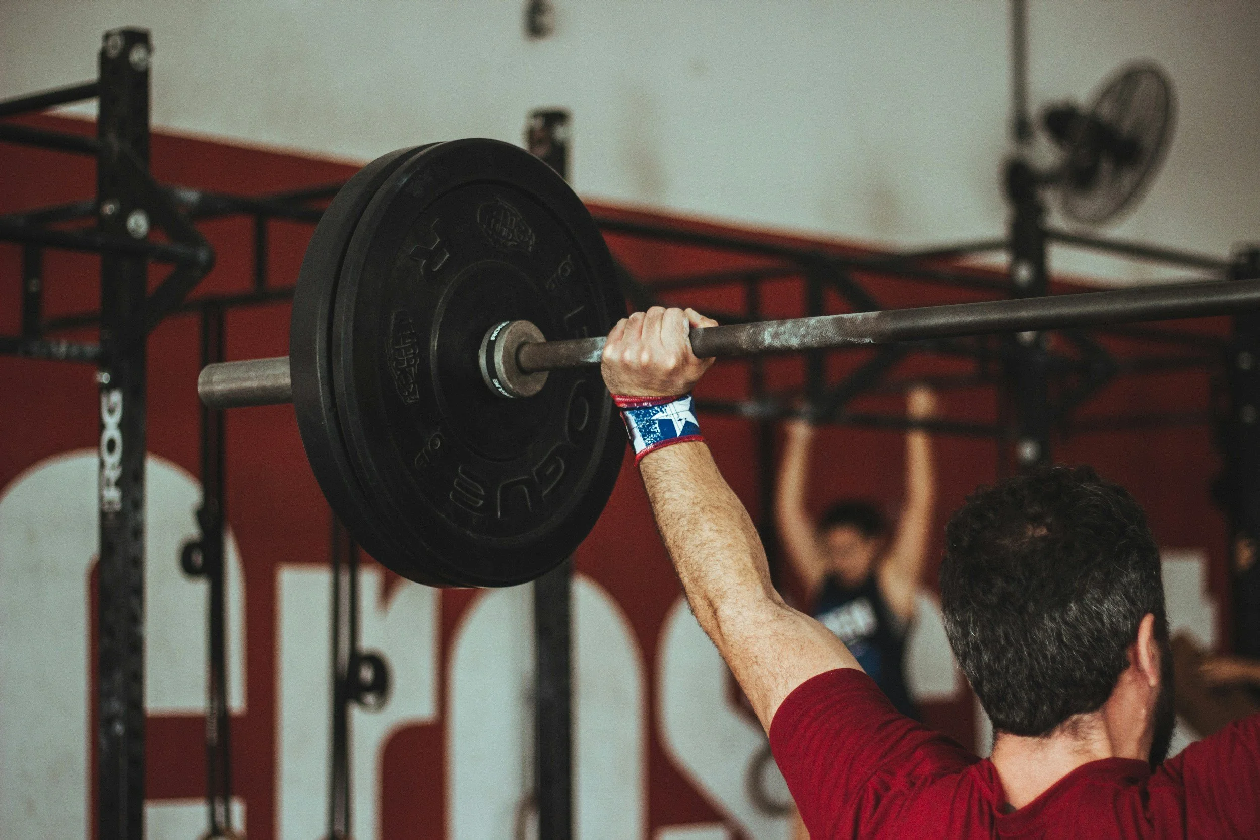 Lifter locking out a barbell overhead in a gym, seen from behind