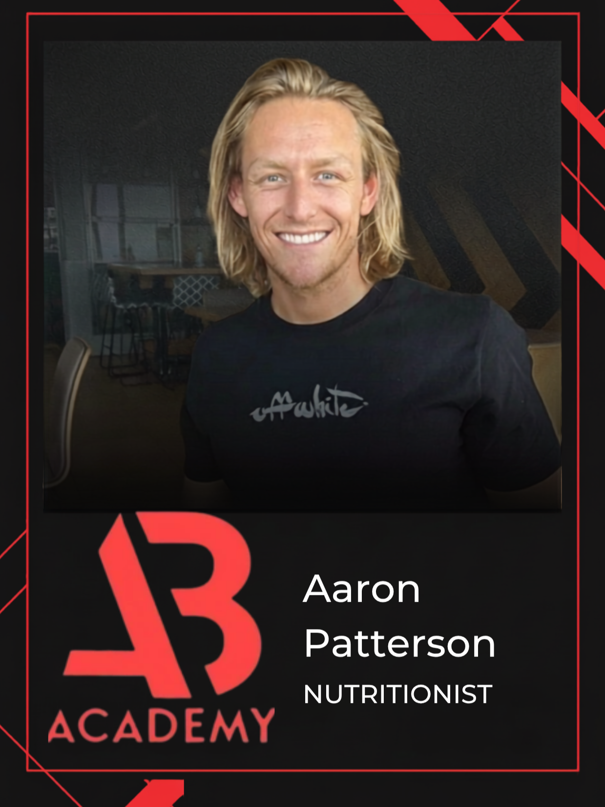 A smiling man with long blonde hair wearing a black t-shirt with white and gray text, sitting indoors with a dark background and a chandelier.
