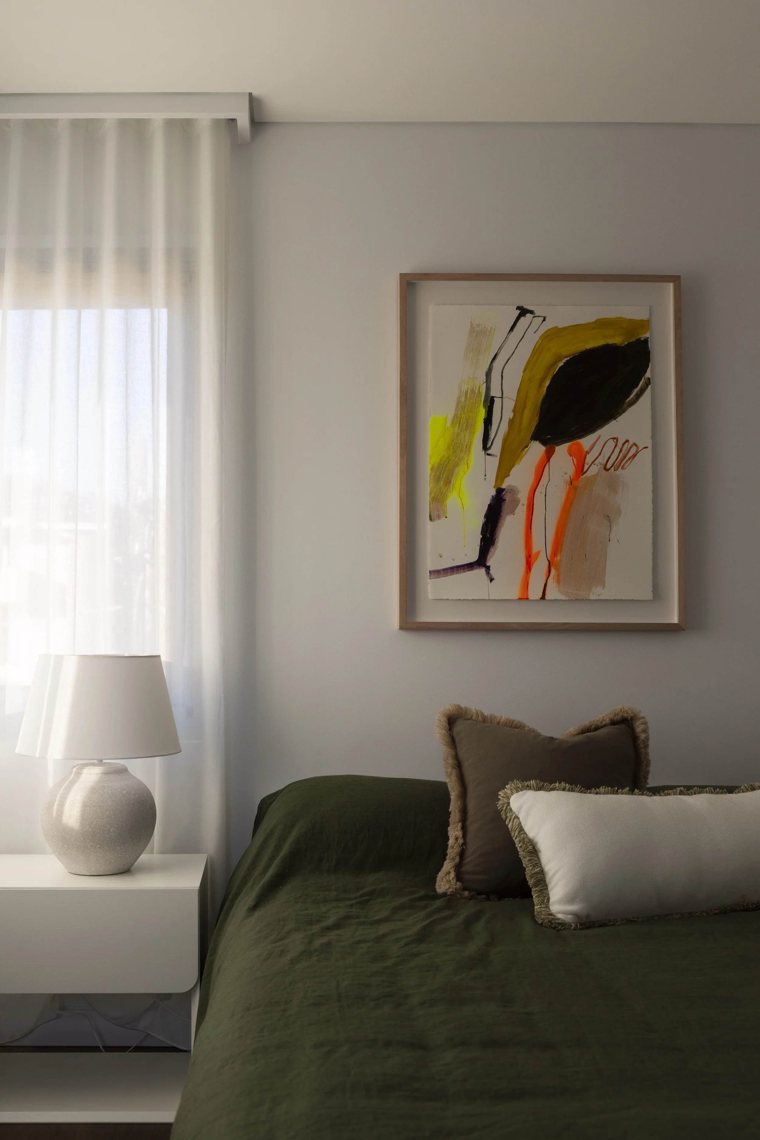 styled bedroom in dover heights with a white nightstand, a white table lamp, a window with sheer curtains, a painting with abstract colorful shapes hanging on the wall, and a bed with dark green bedding and decorative pillows.