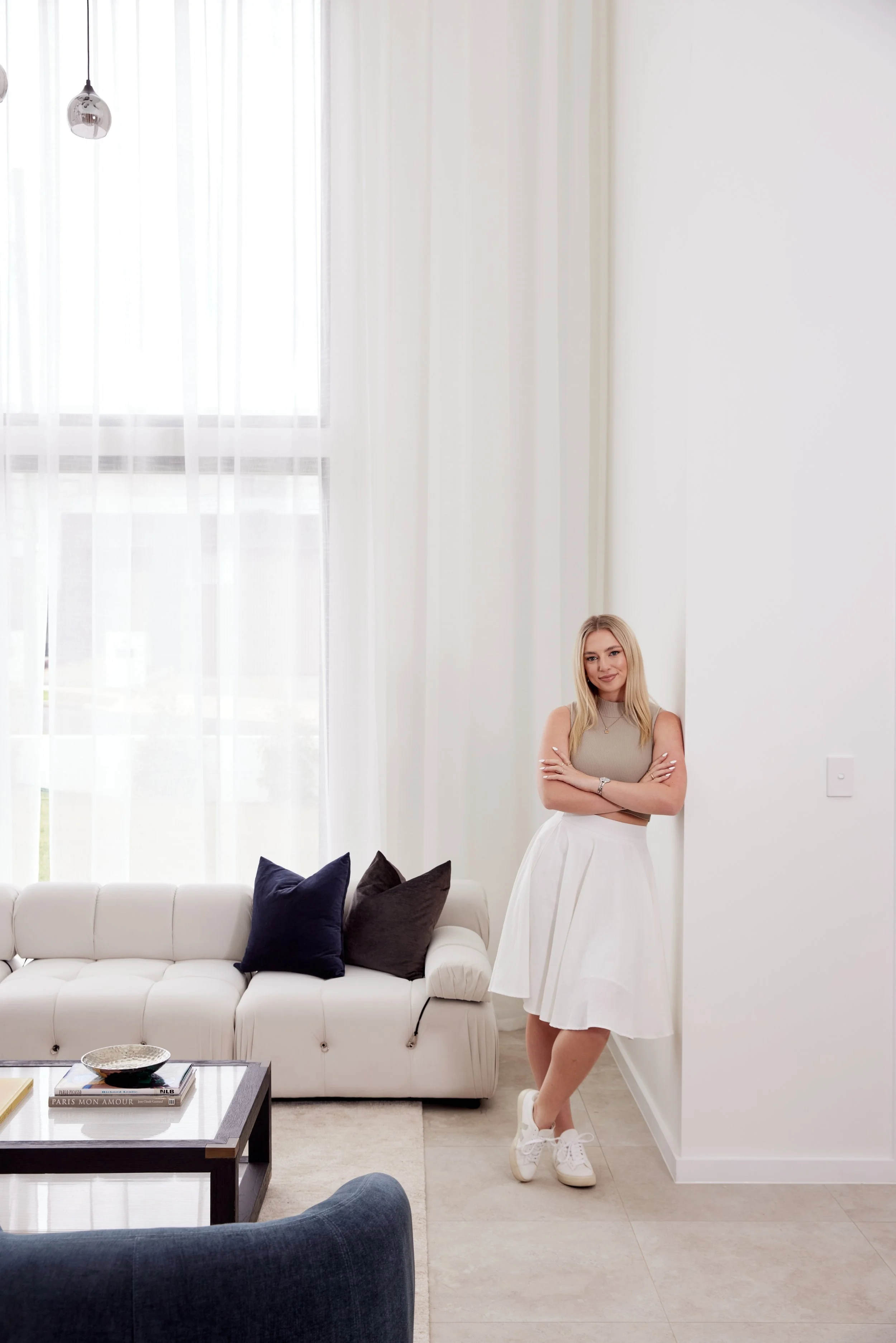 A woman with long blonde hair, wearing a beige sleeveless top, a white pleated skirt, and white sneakers, leaning against a white wall in a bright, modern living room with white curtains, a white sofa with dark pillows, and a glass coffee table.