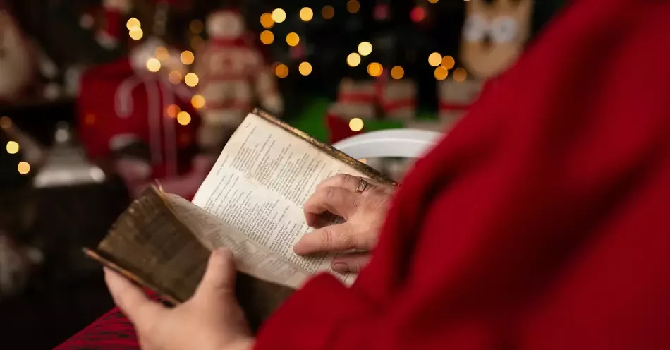 Persona vestida con ropa roja leyendo una Biblia en un entorno decorado con luces navideñas y figuras de Santa Claus.