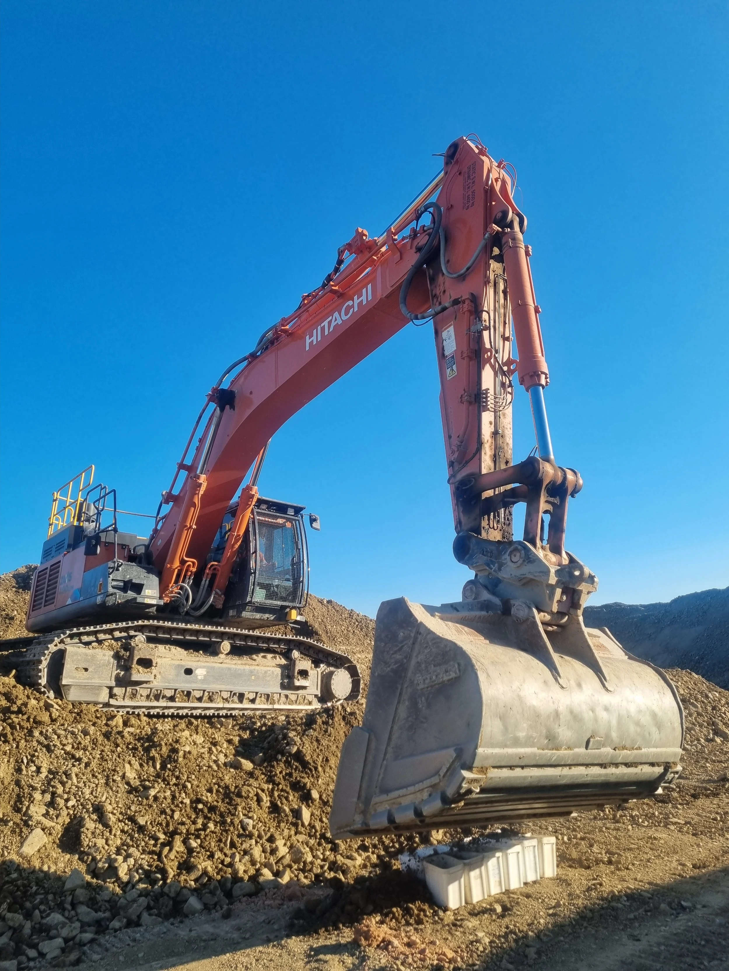 A large orange Hitachi excavator with a bucket attachment digging into dirt against a clear blue sky. Soil testing, buckets