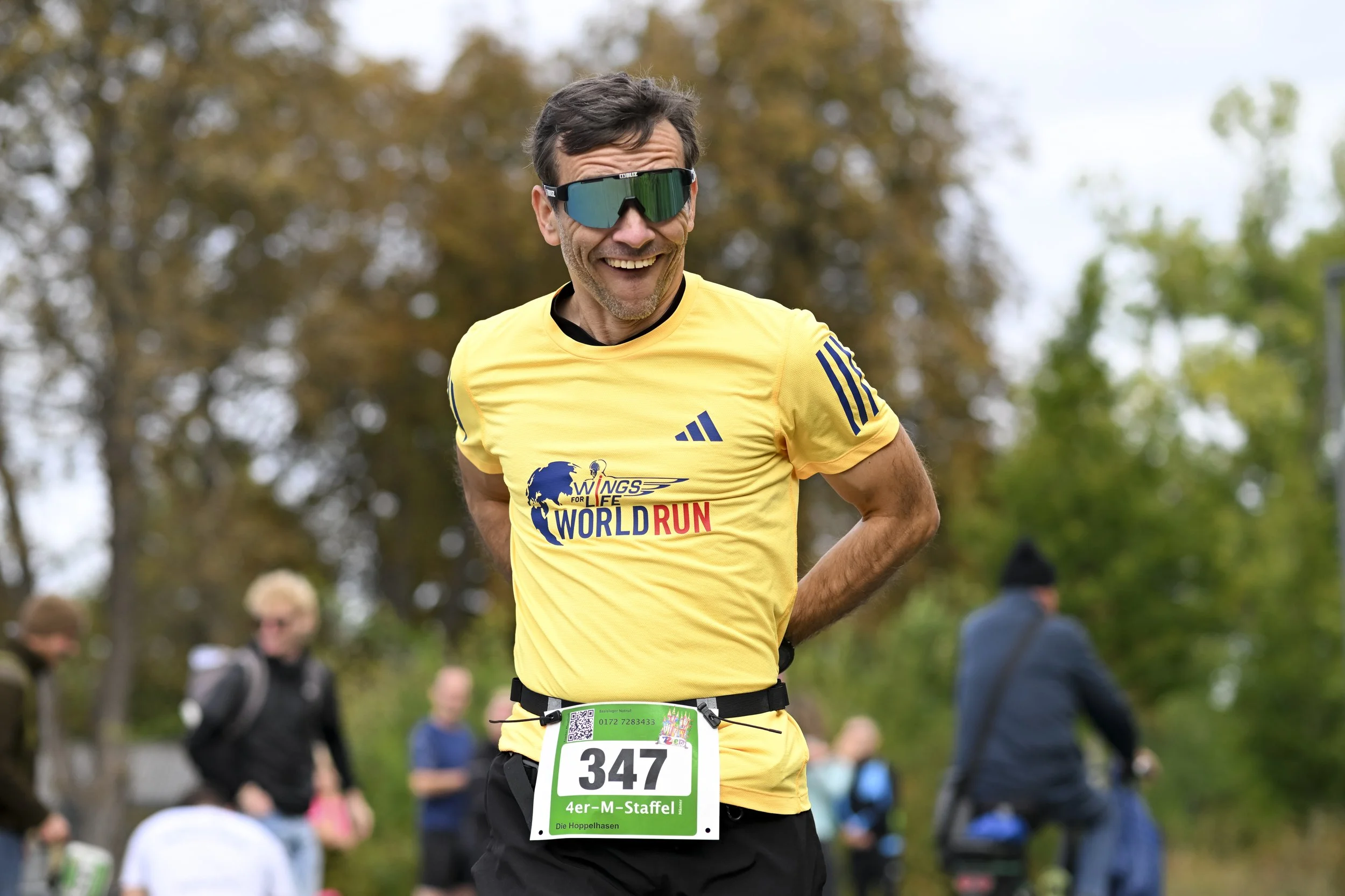 Lächelnder Mann in gelbem Laufshirt mit Race-Nummer 347 bei einem Laufwettbewerb, umgeben von anderen Läufern in einer grünen, herbstlichen Natur.