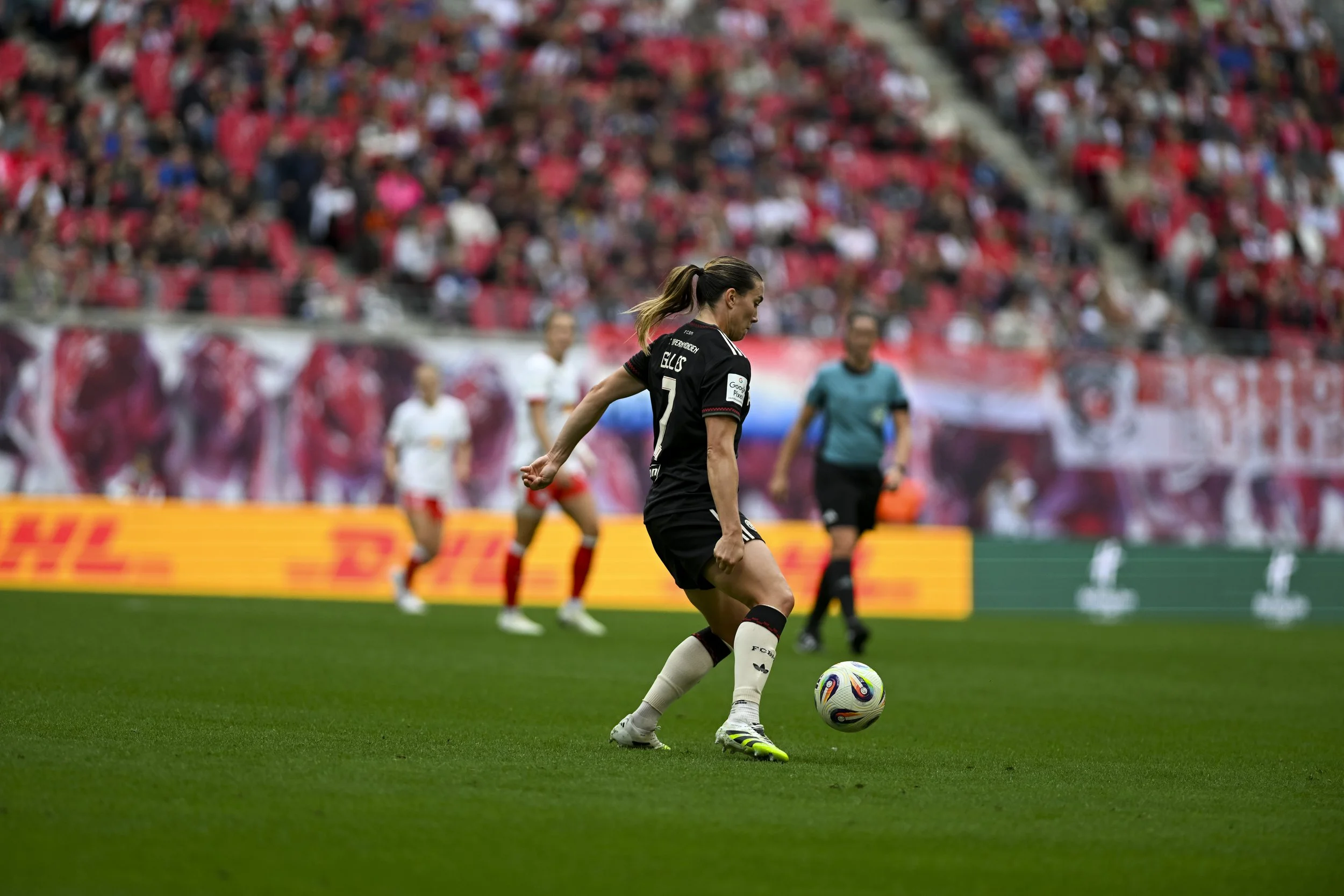 Fußballspielerin in Schwarz bereitet sich vor, den Ball auf dem Spielfeld zu kicken, mit Zuschauer im Hintergrund.