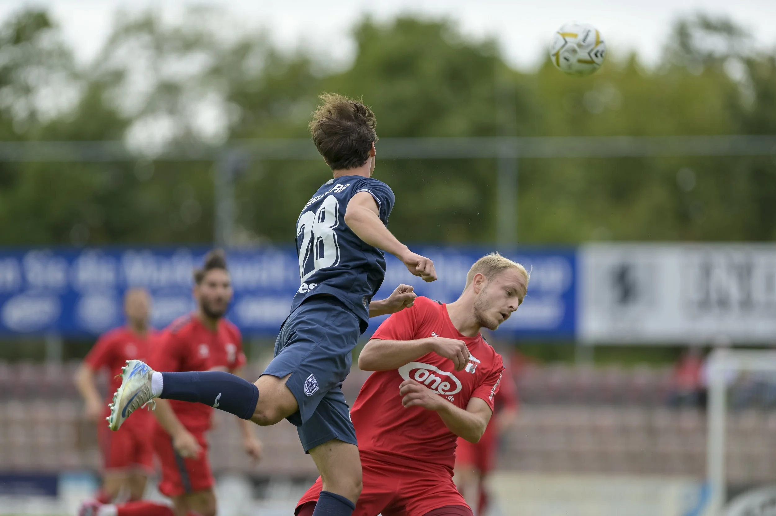 Zwei Fußballspieler, einer in blauer und einer in roter Trikot, kämpfen um den Ball auf dem Spielfeld, im Hintergrund weitere Spieler und Bandenwerbung sichtbar.