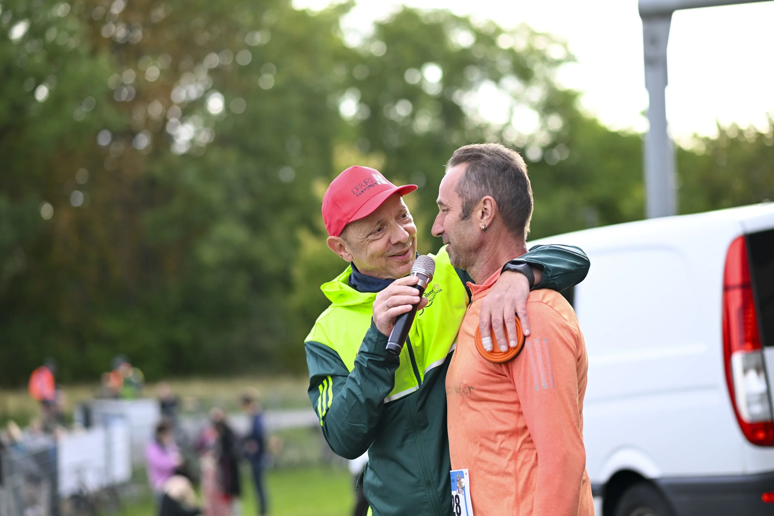 Zwei Männer, einer mit roter Mütze und gelb-grüner Jacke, der andere in orangeener Laufkleidung, stehen sich nahe und sprechen sich freundlich an. Der Mann mit der Mütze hält ein Mikrofon. Im Hintergrund sind mehrere Menschen, Bäume und eine weiße To