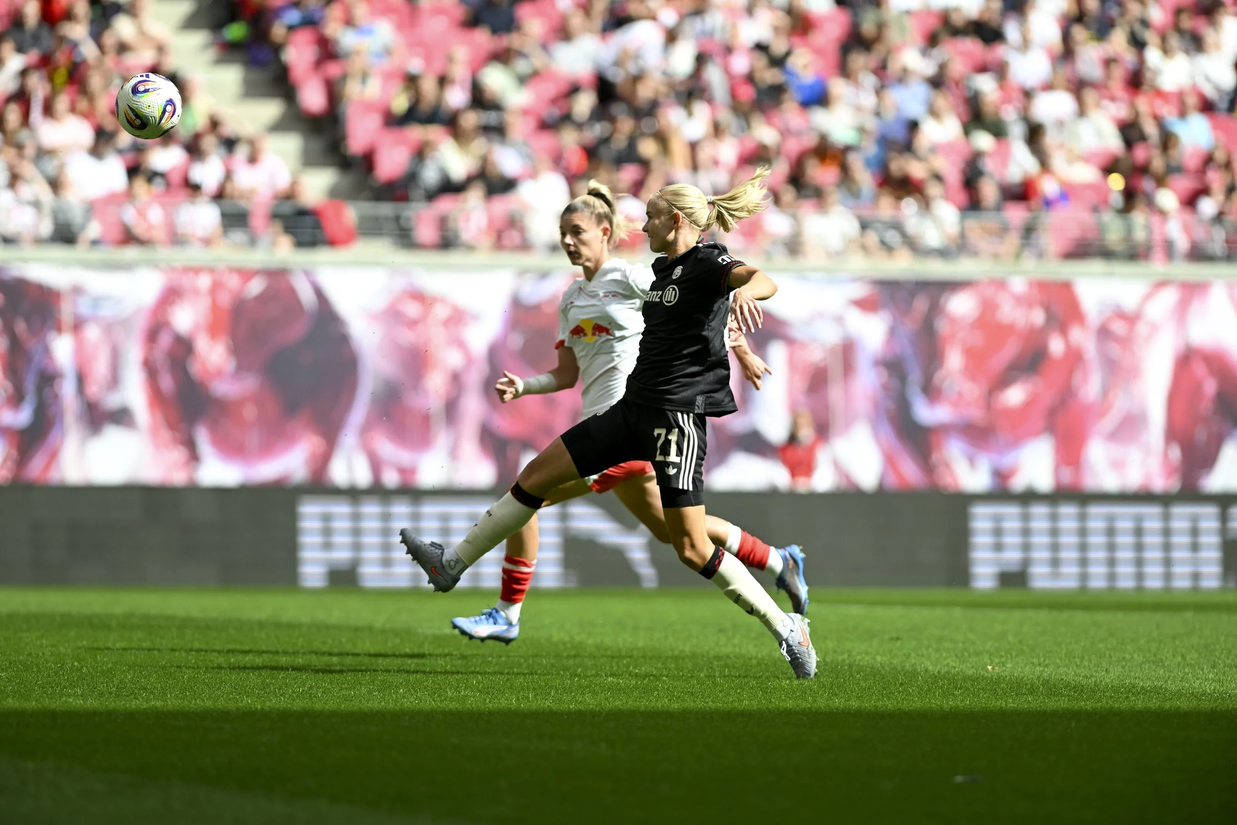 Zwei Fußballspielerinnen im Wettbewerb um den Ball bei einem Spiel, ein Spieler trägt ein schwarzes Trikot mit der Nummer 21, die andere ein weißes Trikot, im Hintergrund ein dicht besetztes Publikum in einem Stadion.