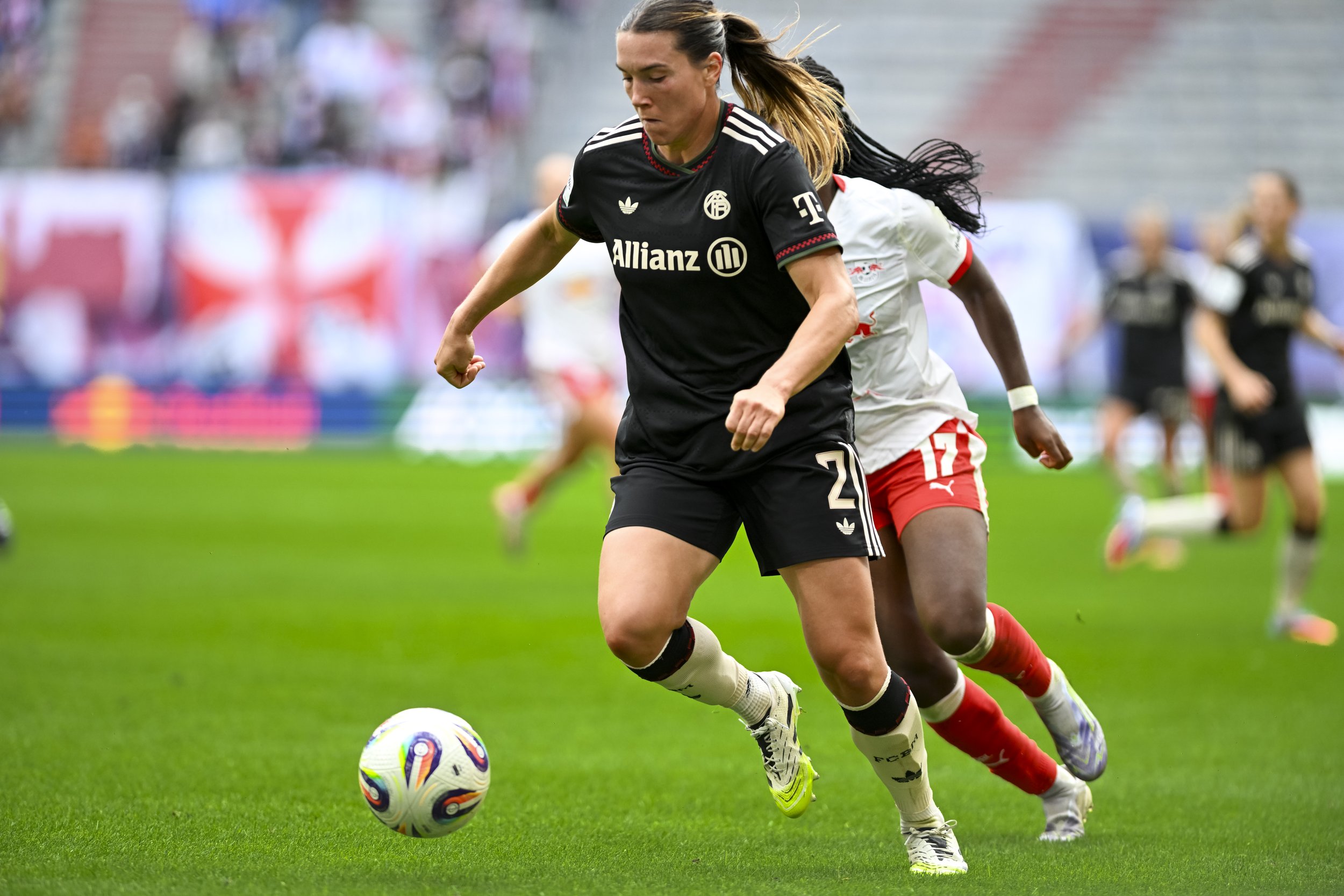 Zwei Fußballspielerinnen kämpfen um den Ball auf dem Spielfeld, eine in schwarzem Trikot, die andere in weiß-rot, im Hintergrund sind Zuschauer im Stadion.
