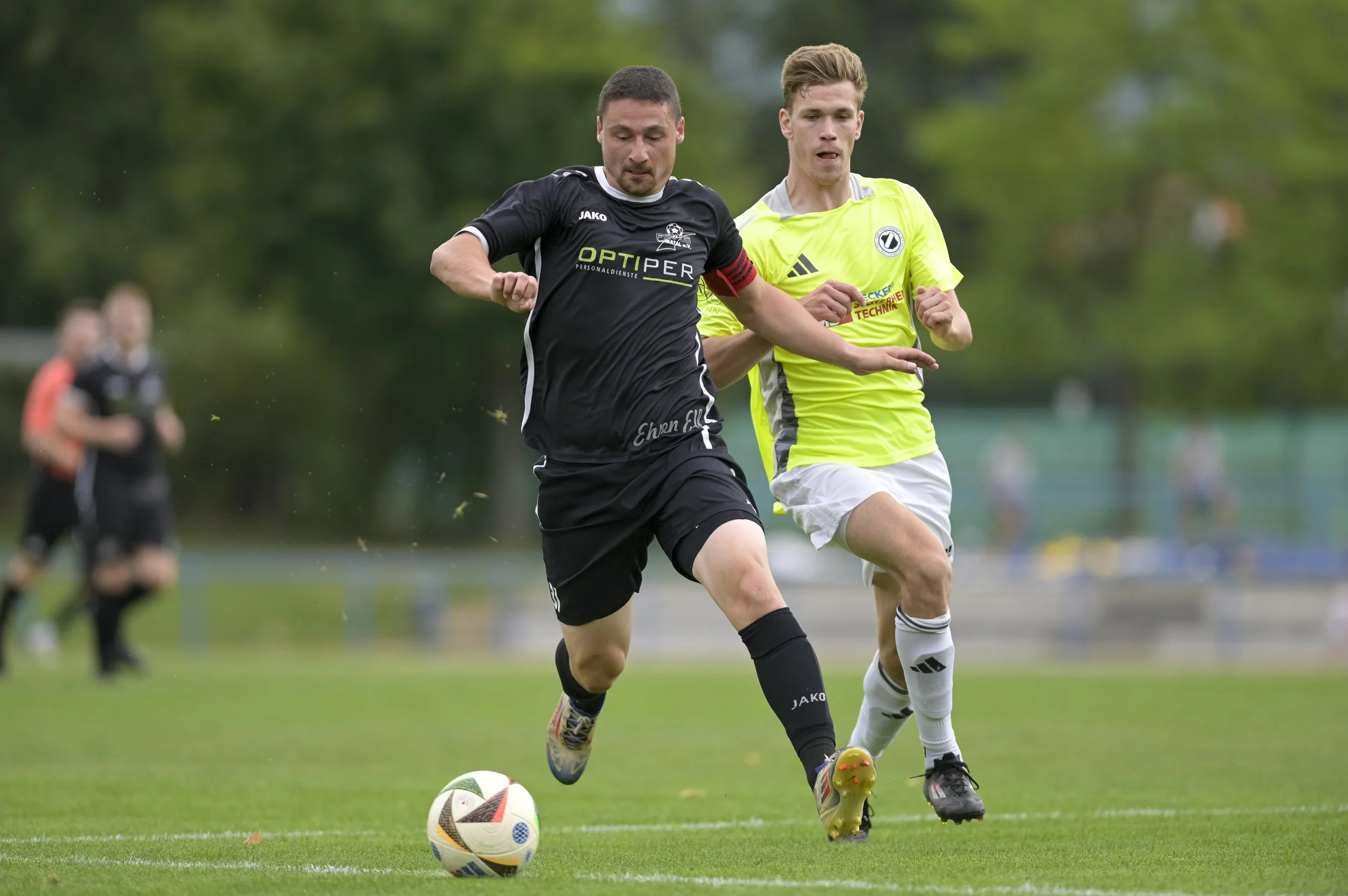 Zwei Fußballspieler kämpfen um den Ball während eines Spiels im Freien, mit weiteren Spielern im Hintergrund.